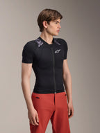 Modelo masculino con la chaqueta protectora Alpinestars A-Motion Plasma de manga corta, color Black Camo con paneles de camuflaje gris en los hombros, protectores integrados en hombros y pecho, logotipo de la estrella blanca en el pecho, tejido de malla transpirable, combinada con pantalones rojos