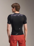 Chaqueta de protección Alpinestars A-Motion Plasma - Manga corta, negra con detalles de camuflaje gris en la parte superior de la espalda, vista trasera que muestra el protector de espalda integrado y logotipo de Astars blanco en el dobladillo, combinada con pantalones cortos rojos, equipo de protección técnica para bicicleta de montaña