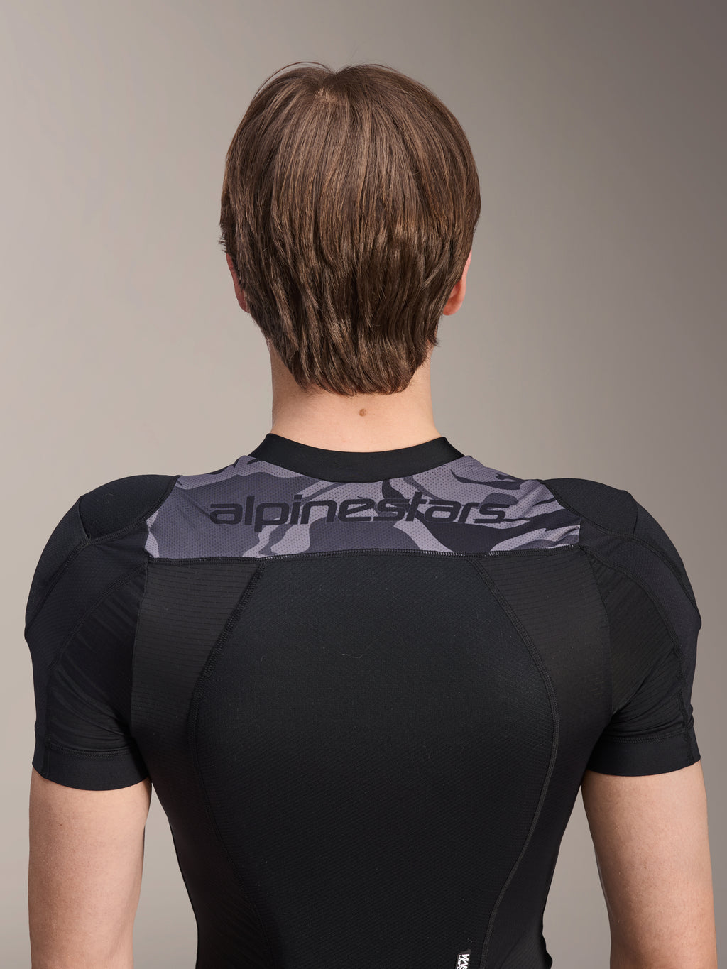 Chaqueta protectora Alpinestars A-Motion Plasma de manga corta, vista trasera de la parte superior del torso, negra con panel de camuflaje gris en los hombros con logotipo de Alpinestars, tejido de malla elástica transpirable para protección de MTB