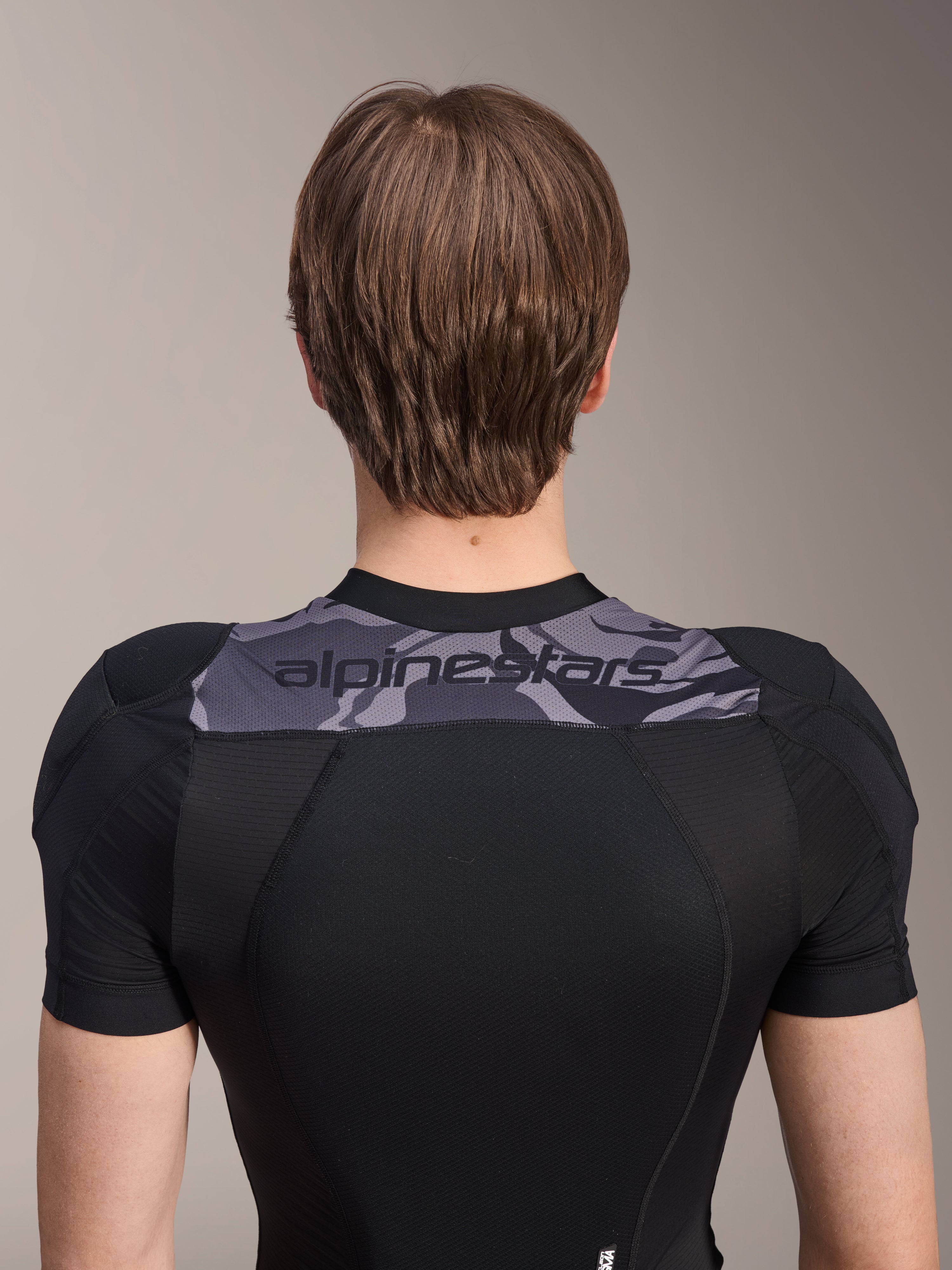 Chaqueta protectora Alpinestars A-Motion Plasma de manga corta, vista trasera de la parte superior del torso, negra con panel de camuflaje gris en los hombros con logotipo de Alpinestars, tejido de malla elástica transpirable para protección de MTB