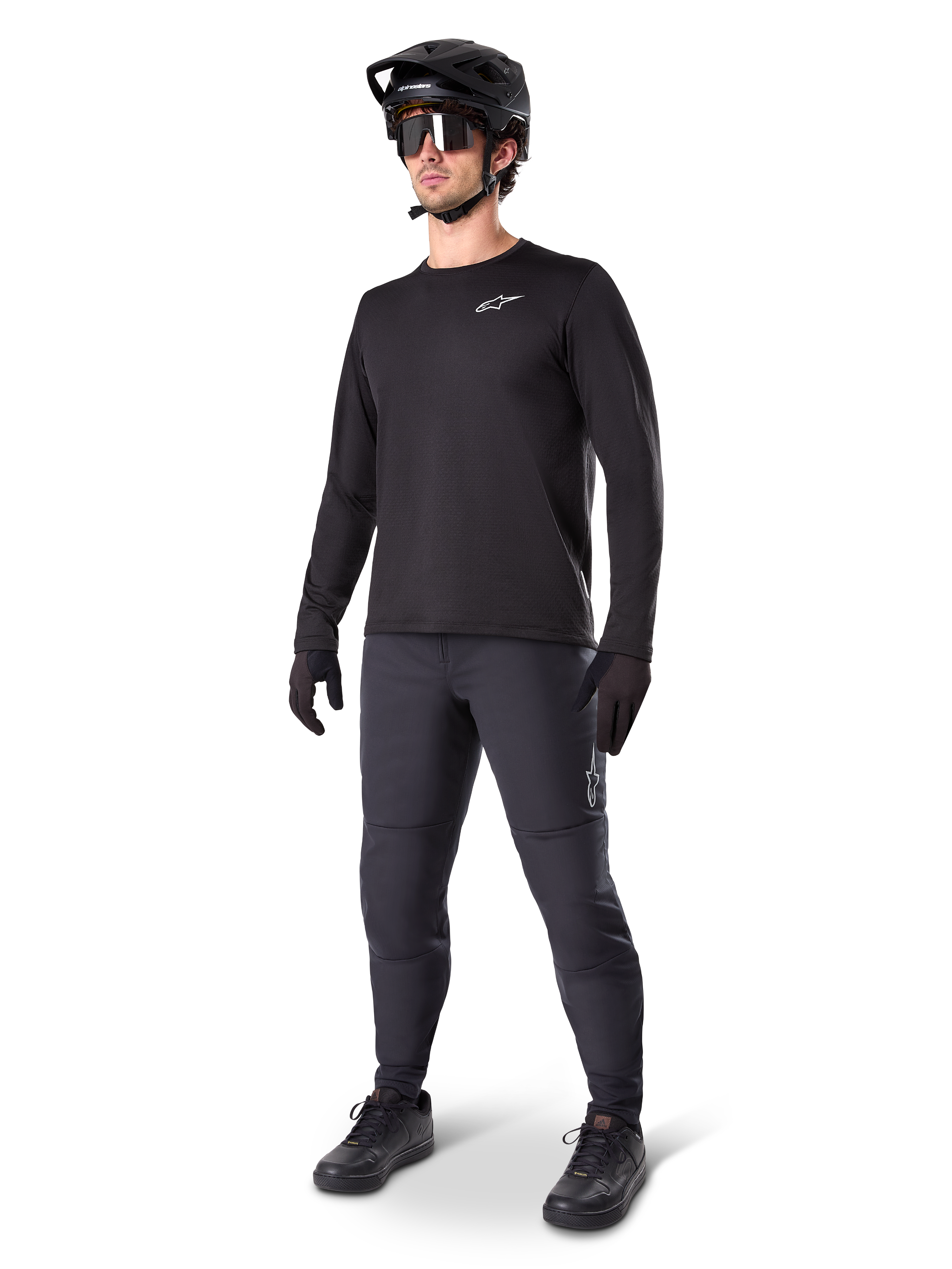 Ciclista de montaña vistiendo los Pantalones Térmicos A-Dura de Alpinestars en color negro, combinados con un jersey negro de manga larga, casco negro y gafas de sol, con el logotipo plateado de Alpinestars en el muslo izquierdo, diseñados para recorrer senderos en climas fríos.