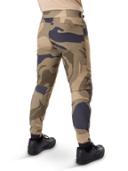 Pantalones Alpinestars A-Dura Camo, pantalones de MTB, estampado de camuflaje marrón con detalles en beige y gris oscuro, vista trasera con zapatillas negras, perneras ajustadas y puños elásticos, tejido elástico en 2 direcciones duradero para ciclismo de montaña
