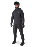 Ciclista de montaña con pantalones impermeables Alpinestars A-Dura en color negro, combinados con una chaqueta impermeable con capucha negra a juego, casco de MTB negro con visera, gafas de ciclismo oscuras y guantes negros, equipo profesional completo para rodar en senderos bajo cualquier condición climática