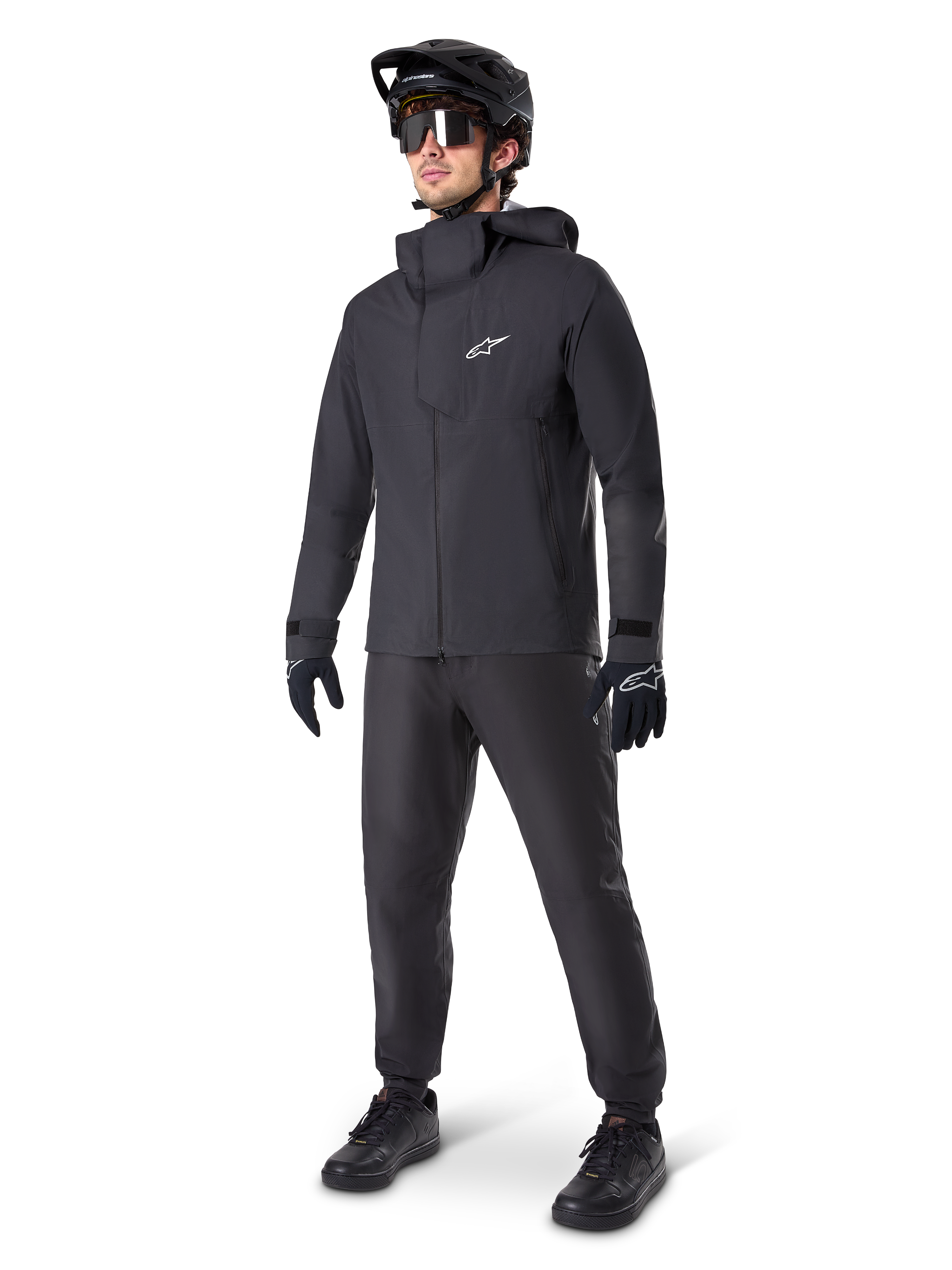 Ciclista de montaña con pantalones impermeables Alpinestars A-Dura en color negro, combinados con una chaqueta impermeable con capucha negra a juego, casco de MTB negro con visera, gafas de ciclismo oscuras y guantes negros, equipo profesional completo para rodar en senderos bajo cualquier condición climática