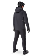 Pantalones Impermeables Alpinestars A-Dura, pantalones de MTB, negro, vistos desde atrás en un ciclista con un conjunto completo de ciclismo de montaña negro que incluye chaqueta con capucha y casco, equipamiento impermeable duradero para senderos embarrados