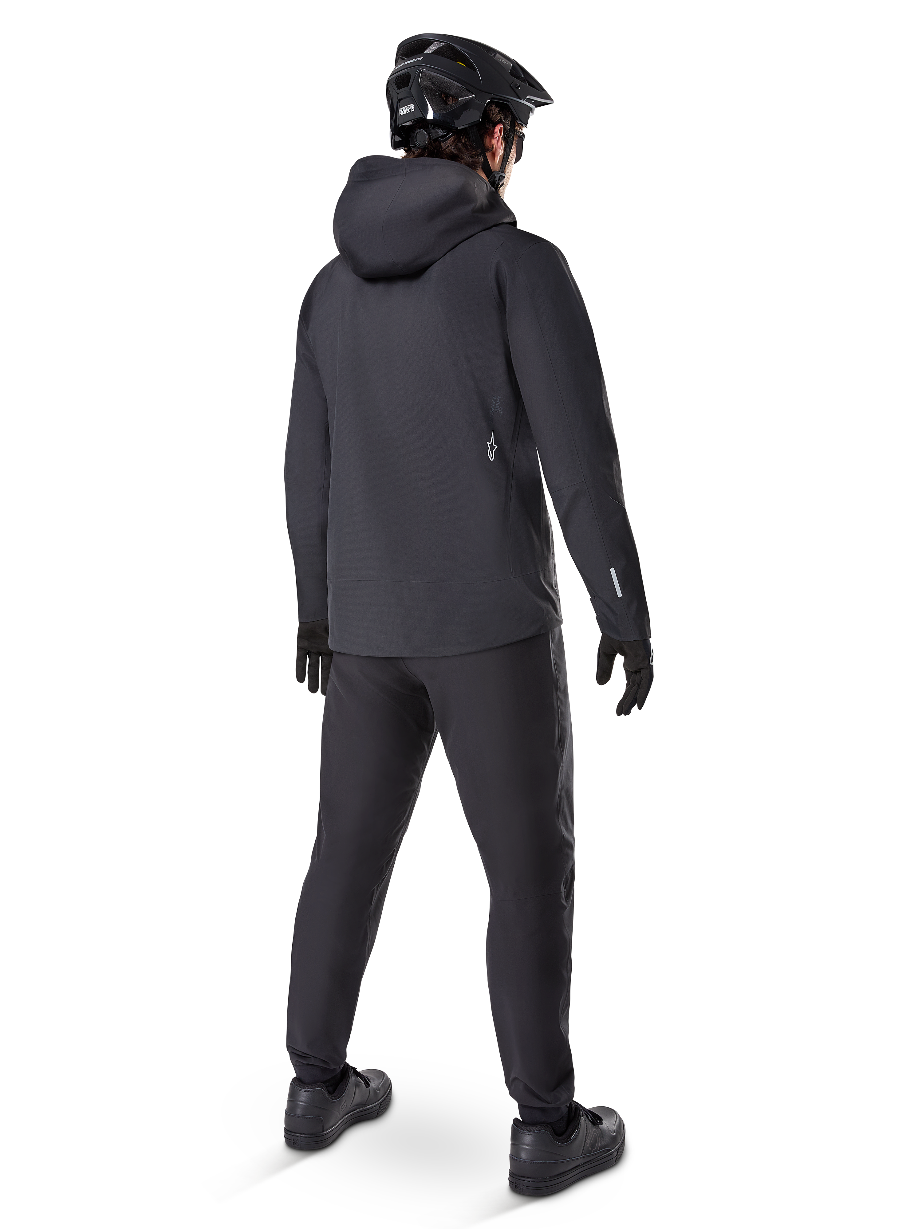 Pantalones Impermeables Alpinestars A-Dura, pantalones de MTB, negro, vistos desde atrás en un ciclista con un conjunto completo de ciclismo de montaña negro que incluye chaqueta con capucha y casco, equipamiento impermeable duradero para senderos embarrados