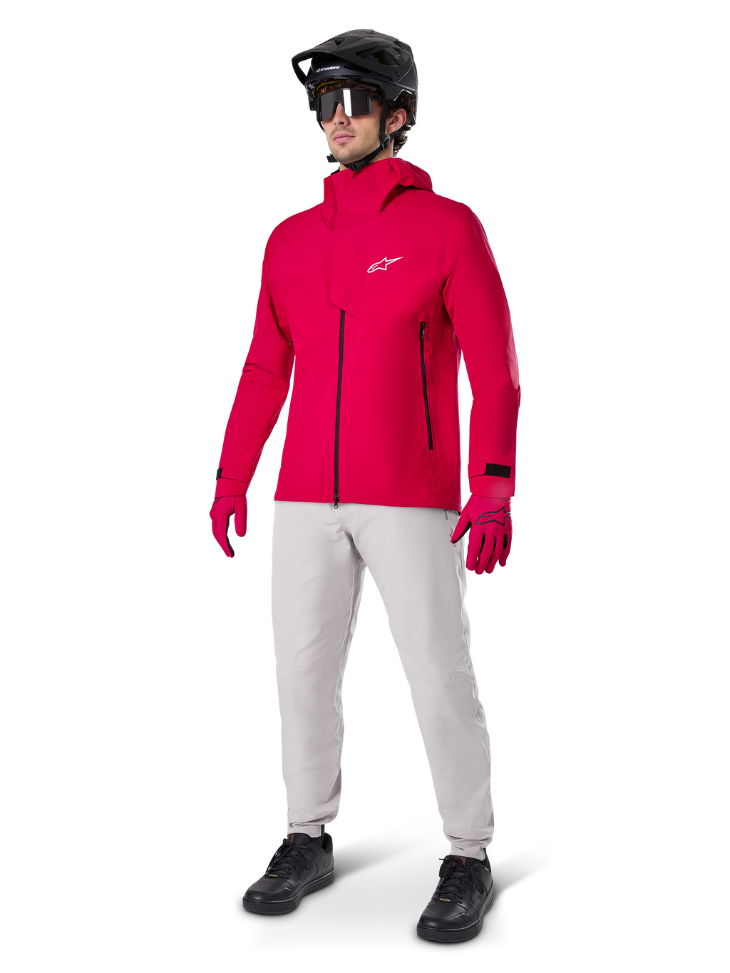 Modelo con pantalones impermeables Alpinestars A-Dura en gris Titanium, combinados con una chaqueta impermeable roja brillante y guantes a juego, casco de bicicleta de montaña negro con gafas y zapatillas negras, equipamiento completo de MTB para condiciones de senderos húmedos