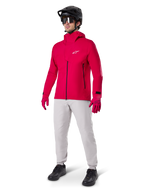 Modelo con pantalones impermeables Alpinestars A-Dura en gris Titanium, combinados con una chaqueta impermeable roja brillante y guantes a juego, casco de bicicleta de montaña negro con gafas y zapatillas negras, equipamiento completo de MTB para condiciones de senderos húmedos