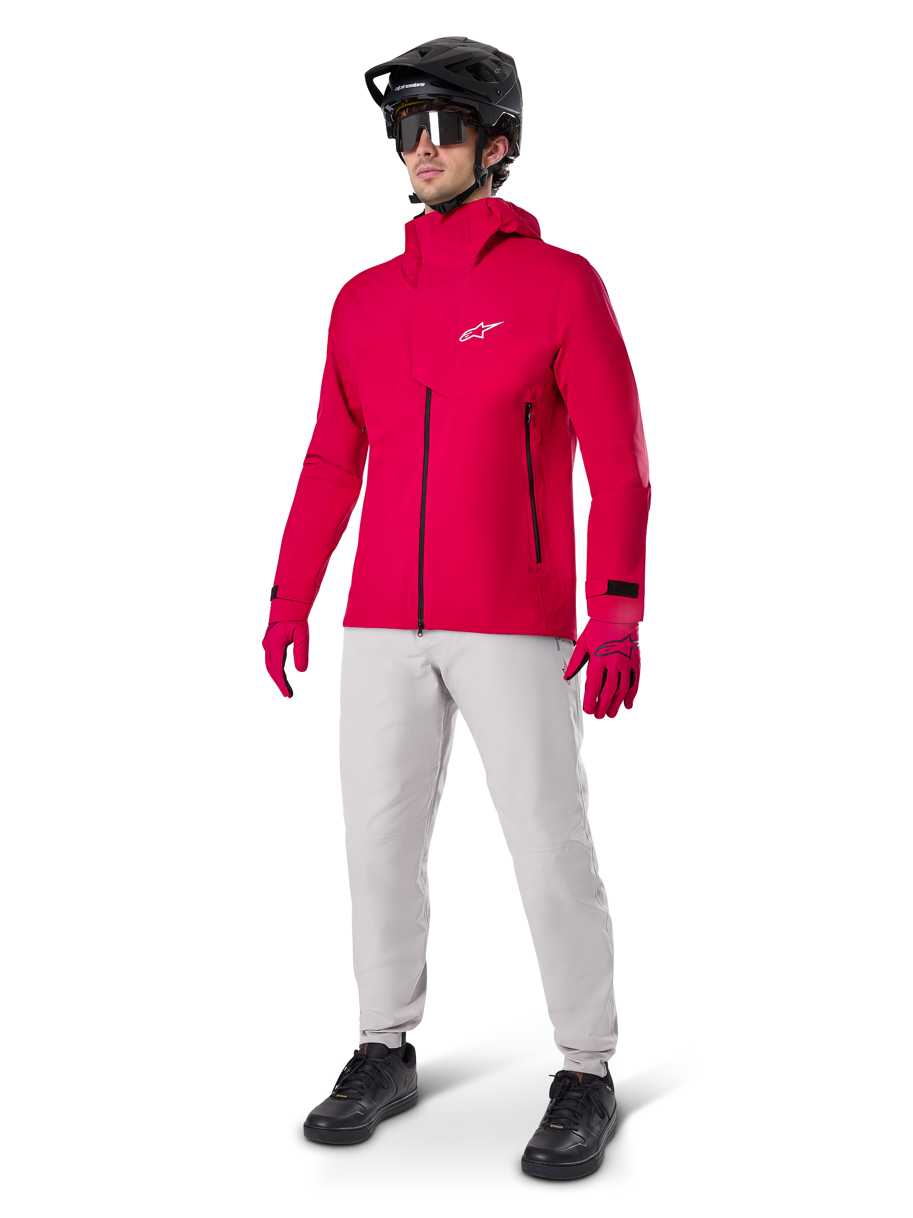 Modelo con pantalones impermeables Alpinestars A-Dura en gris Titanium, combinados con una chaqueta impermeable roja brillante y guantes a juego, casco de bicicleta de montaña negro con gafas y zapatillas negras, equipamiento completo de MTB para condiciones de senderos húmedos