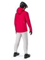 Alpinestars A-Dura Waterproof Pants en color Titanium, vista trasera de un ciclista con los pantalones técnicos para bicicleta de montaña en gris claro, chaqueta roja con capucha y casco negro, equipamiento impermeable y duradero para ciclismo en condiciones húmedas