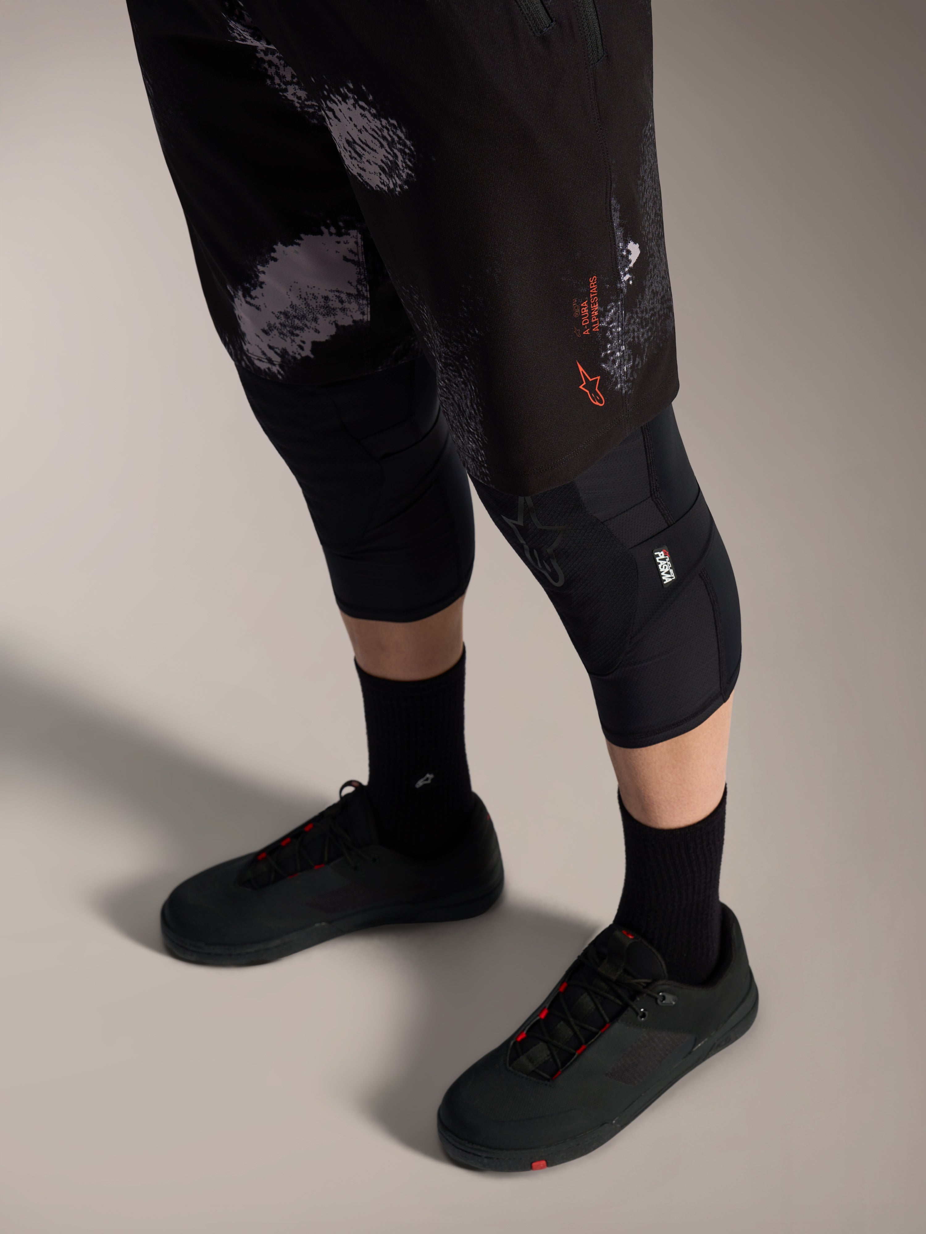 Pantalones cortos Alpinestars A-Dura Ride en Black Camo, equipo MTB con logo naranja, rodilleras y zapatillas.