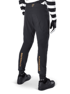 Pantalones Alpinestars A-Aria Elite, pantalones de MTB negros, vista trasera, con la marca Alpinestars en dorado vertical en la parte posterior de las piernas, usados con un jersey a rayas blancas y negras y calzado negro, ropa de ciclismo de alto rendimiento para mountain bike