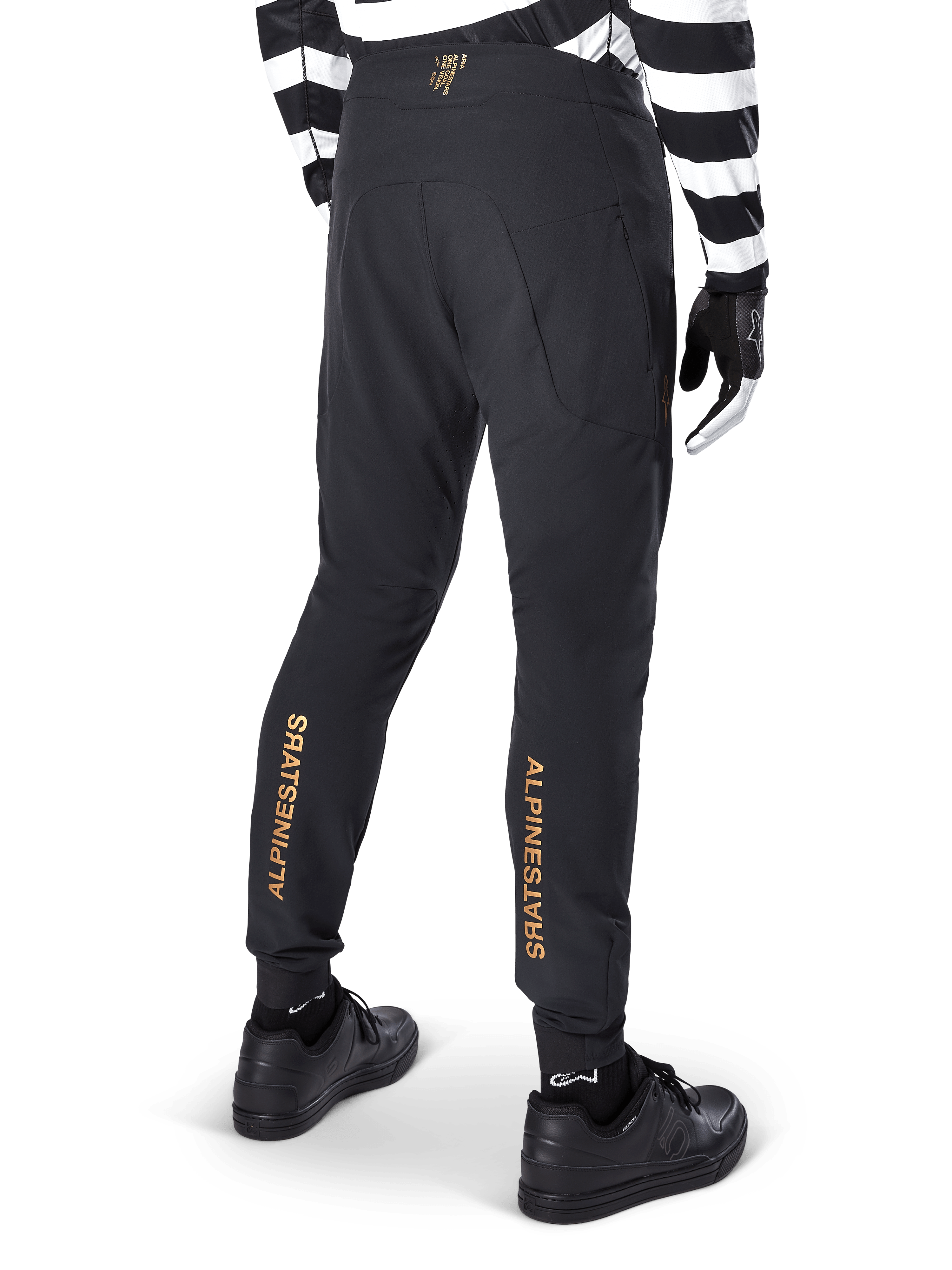 Pantalones Alpinestars A-Aria Elite, pantalones de MTB negros, vista trasera, con la marca Alpinestars en dorado vertical en la parte posterior de las piernas, usados con un jersey a rayas blancas y negras y calzado negro, ropa de ciclismo de alto rendimiento para mountain bike