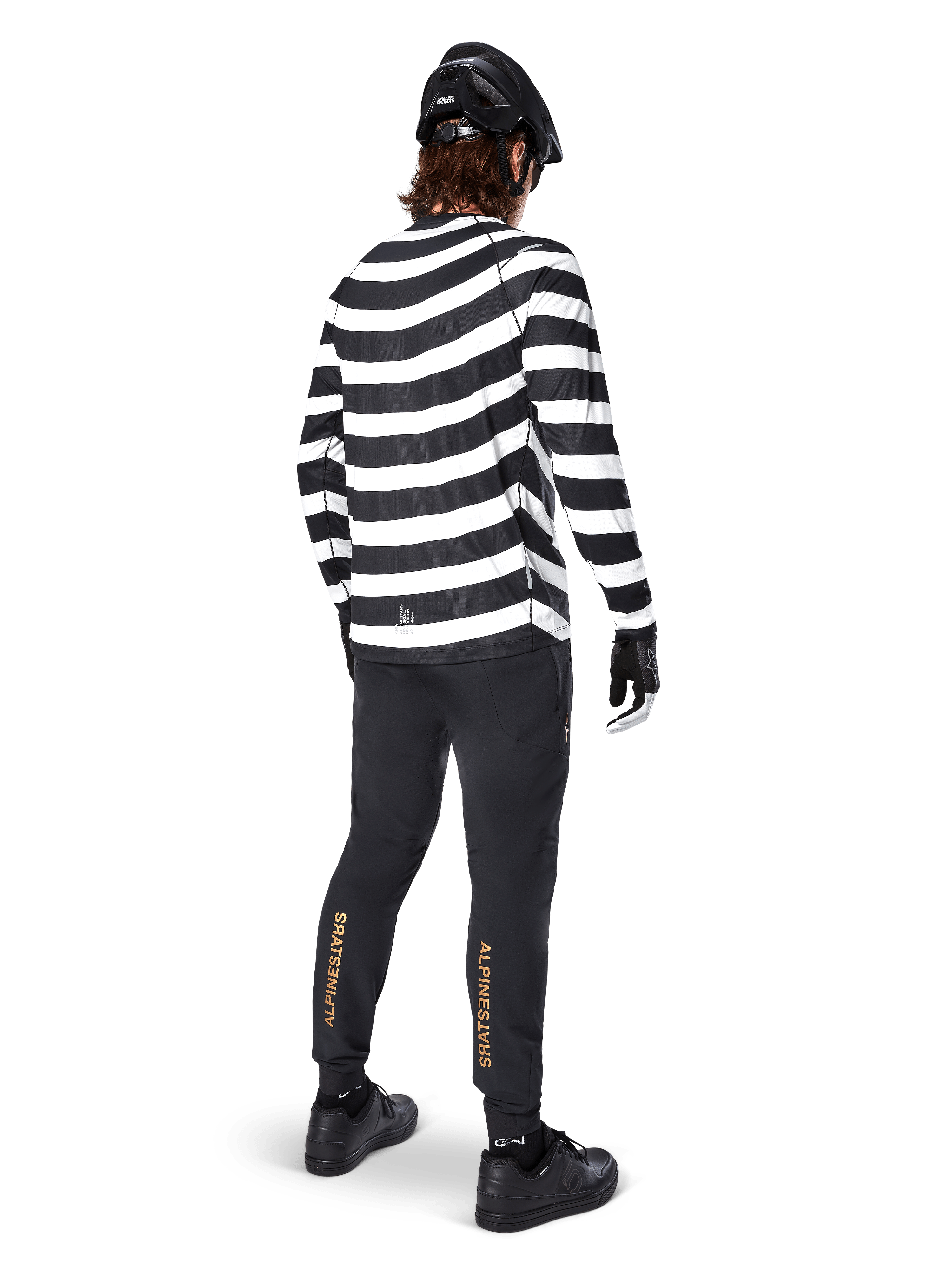 Pantalones Alpinestars A-Aria Elite, pantalones de MTB negros con un logotipo vertical dorado en las pantorrillas, usados por un ciclista con un jersey de manga larga a rayas blancas y negras, casco negro y zapatillas negras, vista posterior que muestra el ajuste de rendimiento y el diseño para senderos