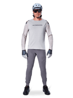 Pantalones Alpinestars A-Aria Elite, pantalones de ciclismo de montaña gris oscuro, usados por un ciclista con jersey de manga larga gris claro, casco azul claro y calzado negro, con perforaciones láser visibles en la parte interna de las piernas y un ajuste de rendimiento entallado