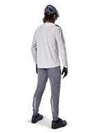 Pantalones Alpinestars A-Aria Elite, pantalones de ciclismo MTB, gris oscuro, vista trasera de un ciclista con la equipación completa, con logotipos blancos de Alpinestars en la parte inferior de las piernas, ajuste de rendimiento con perforaciones láser, combinados con un jersey de manga larga gris claro y casco de MTB gris