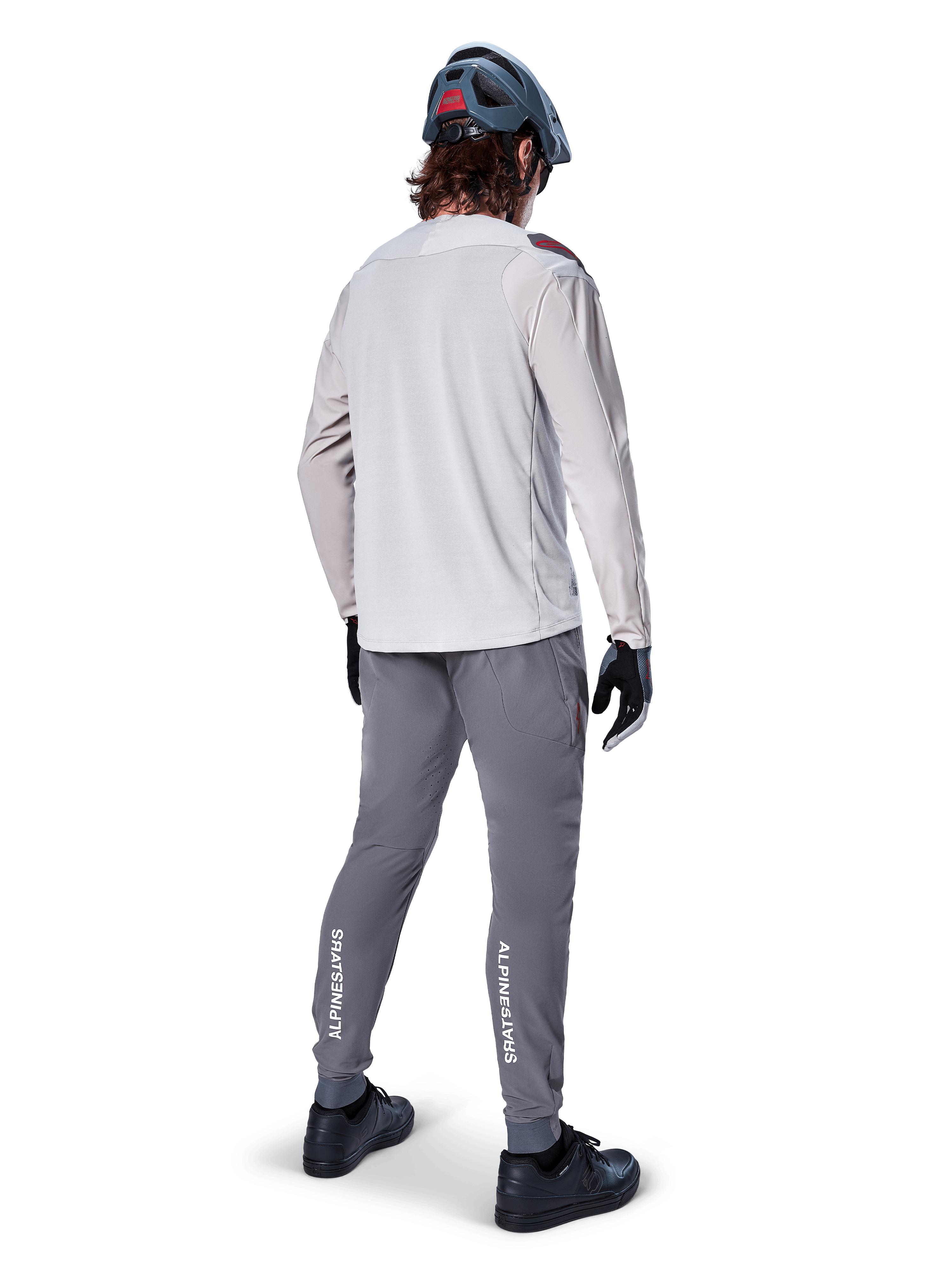 Pantalones Alpinestars A-Aria Elite, pantalones de ciclismo MTB, gris oscuro, vista trasera de un ciclista con la equipación completa, con logotipos blancos de Alpinestars en la parte inferior de las piernas, ajuste de rendimiento con perforaciones láser, combinados con un jersey de manga larga gris claro y casco de MTB gris