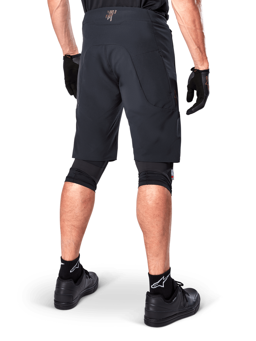 Vista trasera de un ciclista con los shorts Alpinestars A-Aria Elite en color negro, pantalones cortos de alto rendimiento para bicicleta de montaña, combinados con guantes de ciclismo negros y zapatillas negras, diseñados para ofrecer flexibilidad de movimiento en senderos de montaña