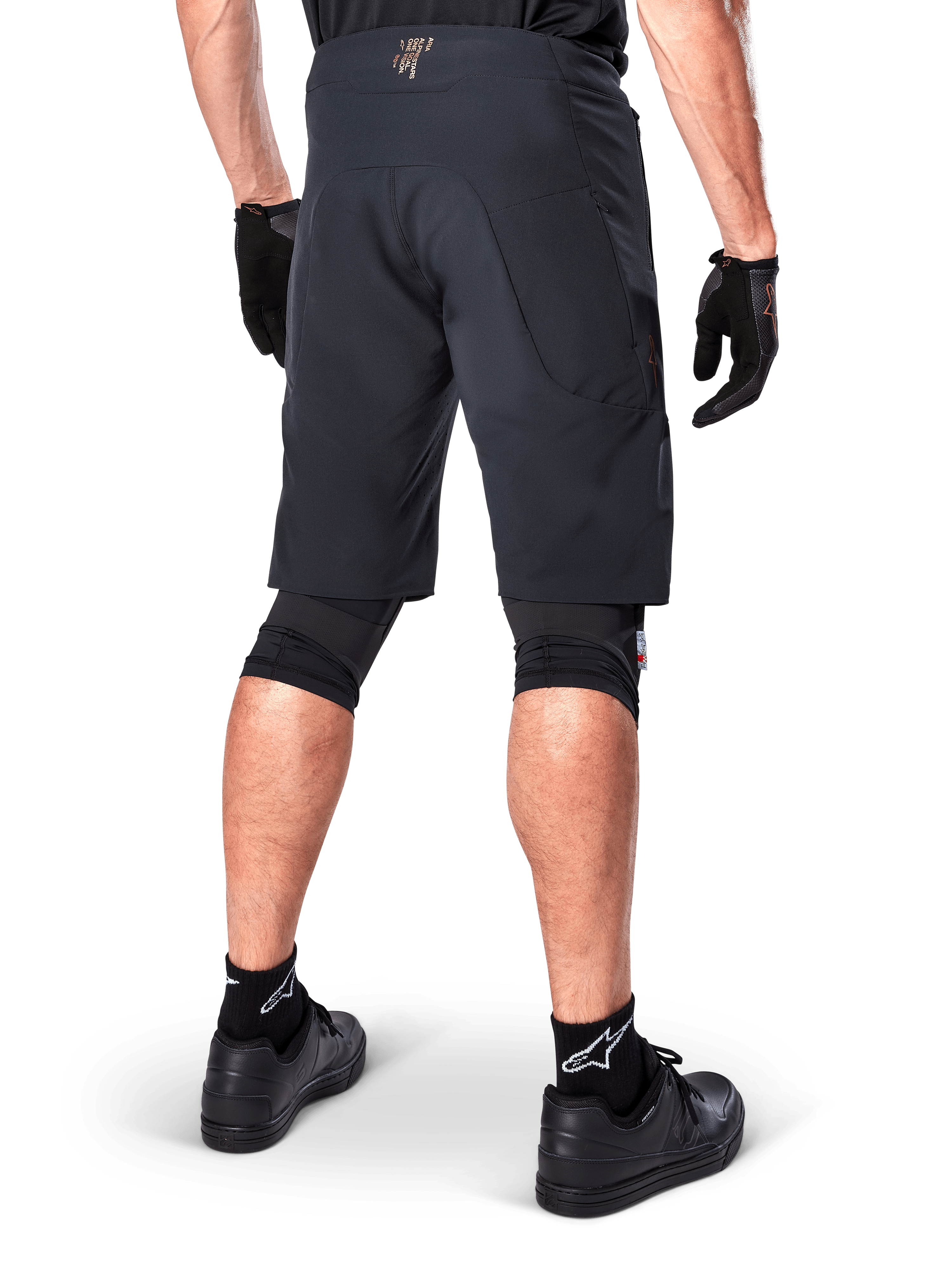 Vista trasera de un ciclista con los shorts Alpinestars A-Aria Elite en color negro, pantalones cortos de alto rendimiento para bicicleta de montaña, combinados con guantes de ciclismo negros y zapatillas negras, diseñados para ofrecer flexibilidad de movimiento en senderos de montaña