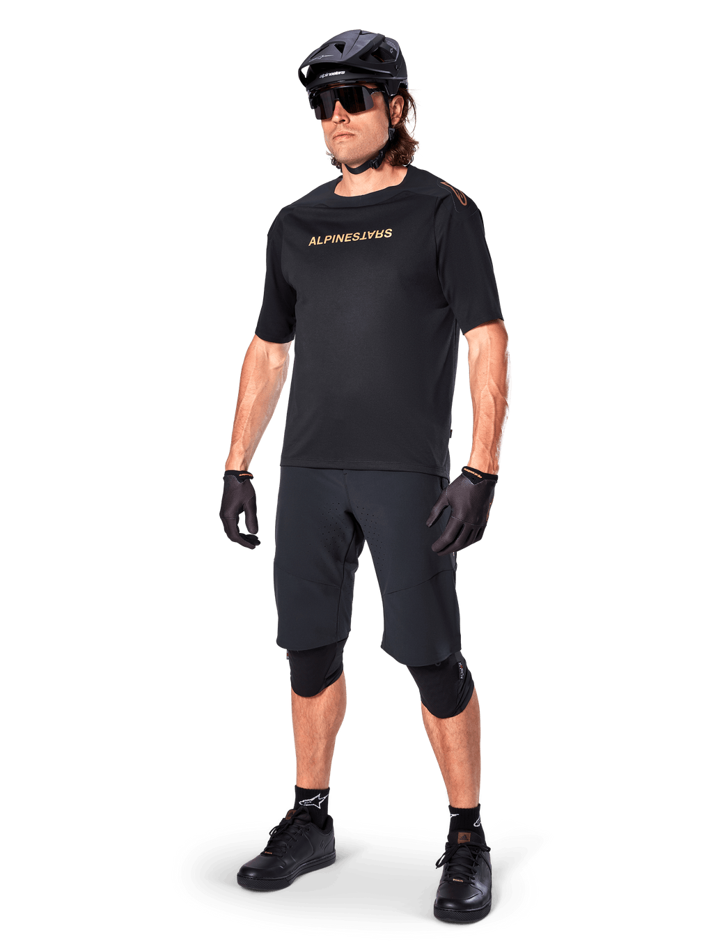 Ciclista con los shorts Alpinestars A-Aria Elite, pantalones cortos negros para bicicleta de montaña, combinados con un jersey negro a juego con logotipos dorados, casco negro, gafas de sol y rodilleras, equipamiento completo de alto rendimiento para ciclismo de montaña
