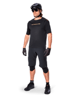 Ciclista con los shorts Alpinestars A-Aria Elite, pantalones cortos negros para bicicleta de montaña, combinados con un jersey negro a juego con logotipos dorados, casco negro, gafas de sol y rodilleras, equipamiento completo de alto rendimiento para ciclismo de montaña