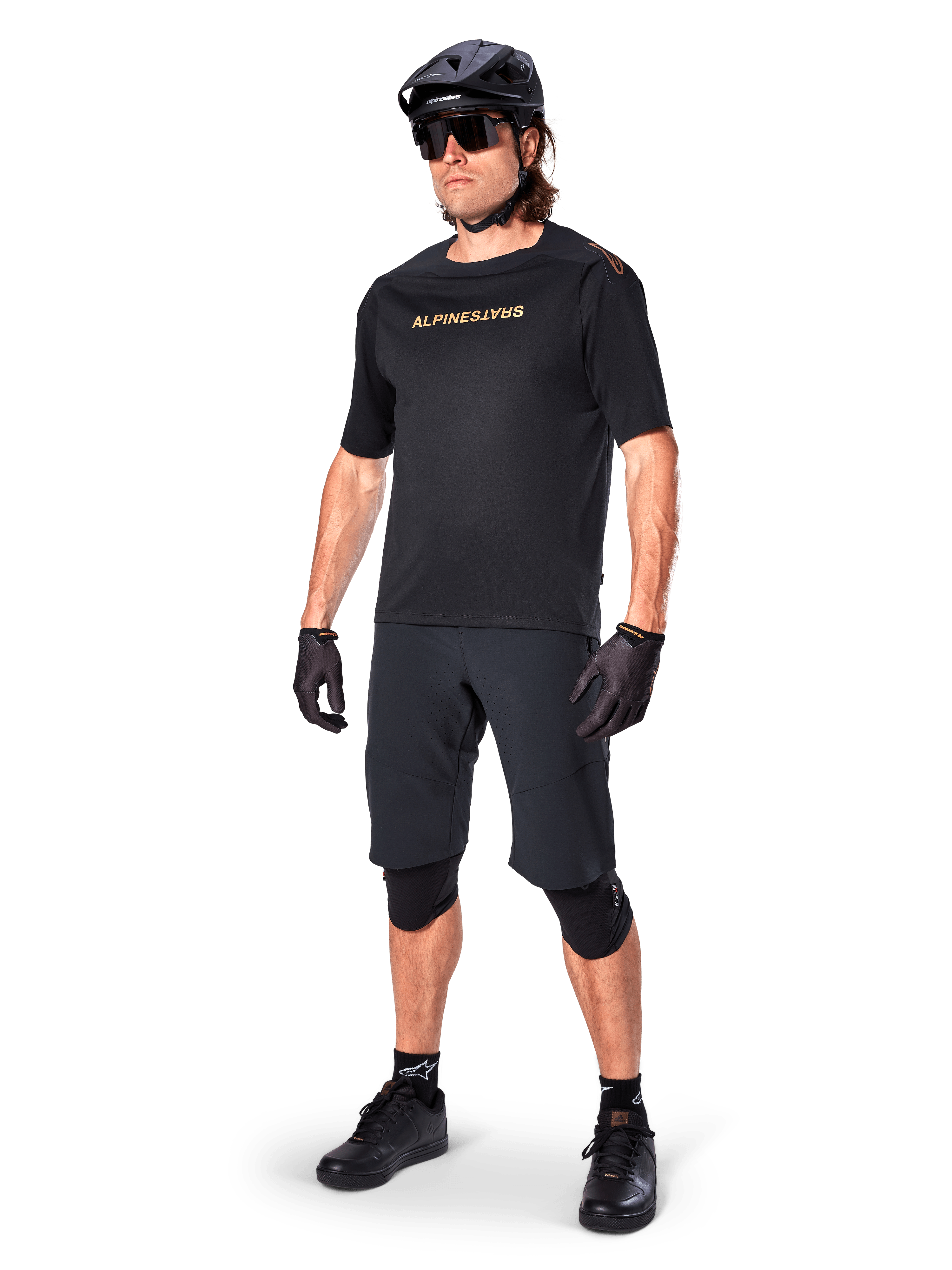 Ciclista con los shorts Alpinestars A-Aria Elite, pantalones cortos negros para bicicleta de montaña, combinados con un jersey negro a juego con logotipos dorados, casco negro, gafas de sol y rodilleras, equipamiento completo de alto rendimiento para ciclismo de montaña