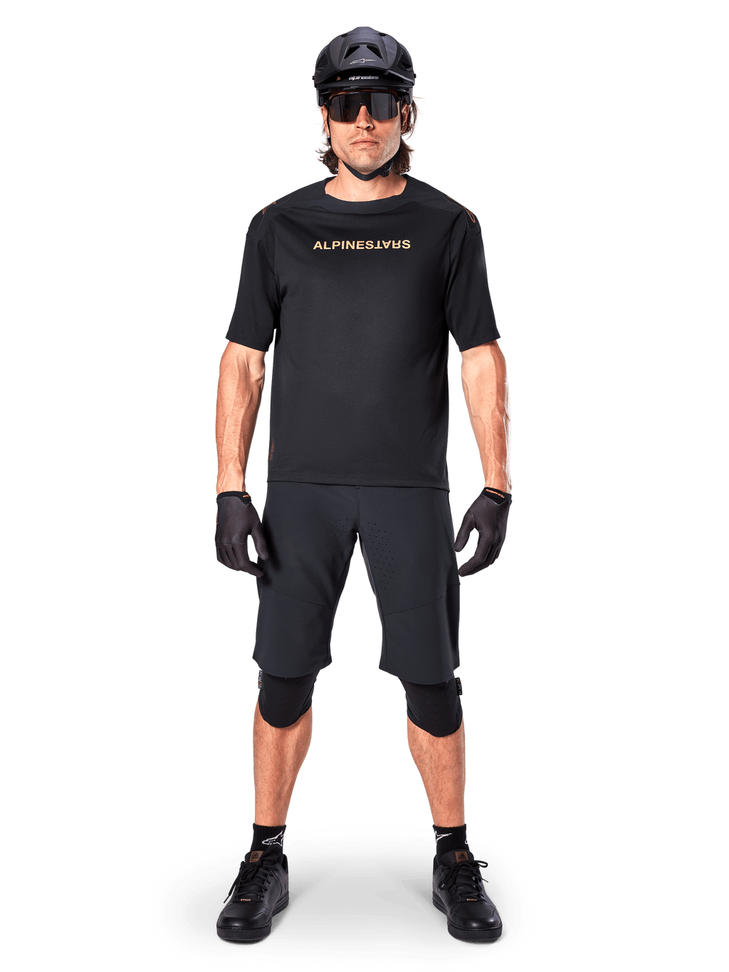 Ciclista de montaña masculino con shorts Alpinestars A-Aria Elite en color negro, combinados con un jersey de manga corta Alpinestars negro a juego, casco, guantes y rodilleras negros, conjunto completo de equipamiento para ciclismo todoterreno