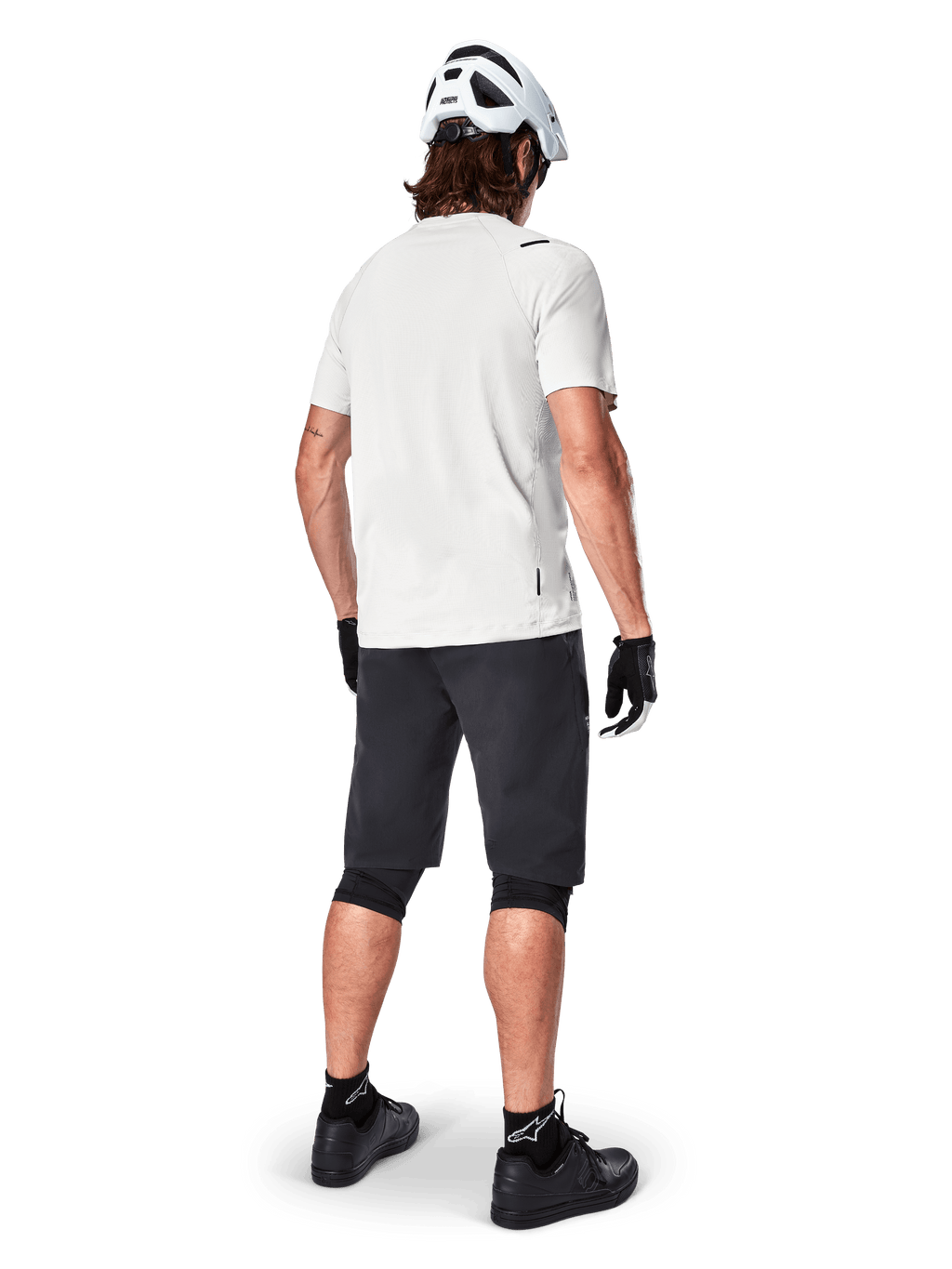 Ciclista de montaña visto desde atrás con shorts Alpinestars A-Aria en color negro, jersey blanco de manga corta de alto rendimiento, casco blanco, guantes negros y zapatillas de ciclismo de montaña negras, equipamiento de ciclismo para trail