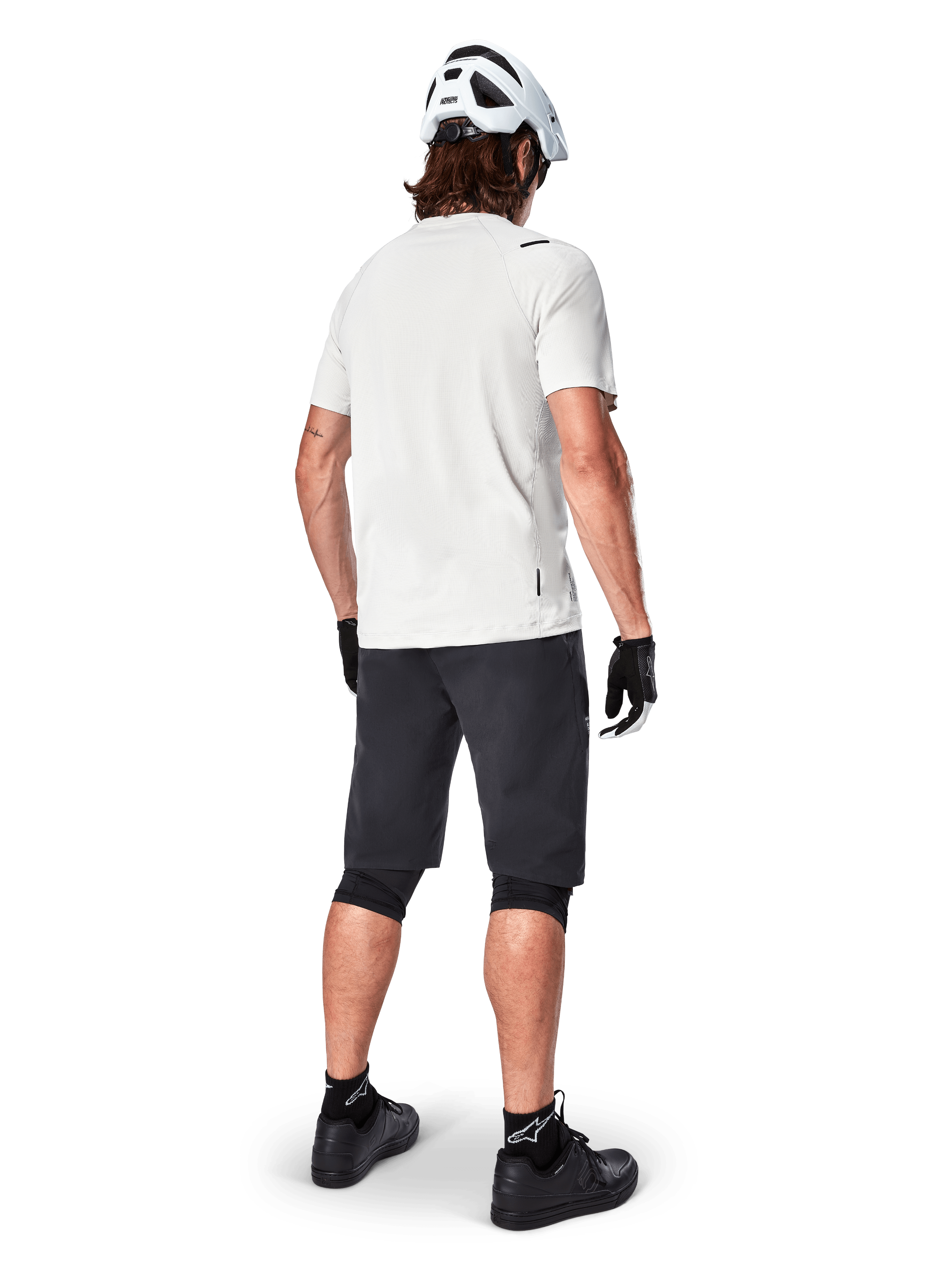 Ciclista de montaña visto desde atrás con shorts Alpinestars A-Aria en color negro, jersey blanco de manga corta de alto rendimiento, casco blanco, guantes negros y zapatillas de ciclismo de montaña negras, equipamiento de ciclismo para trail