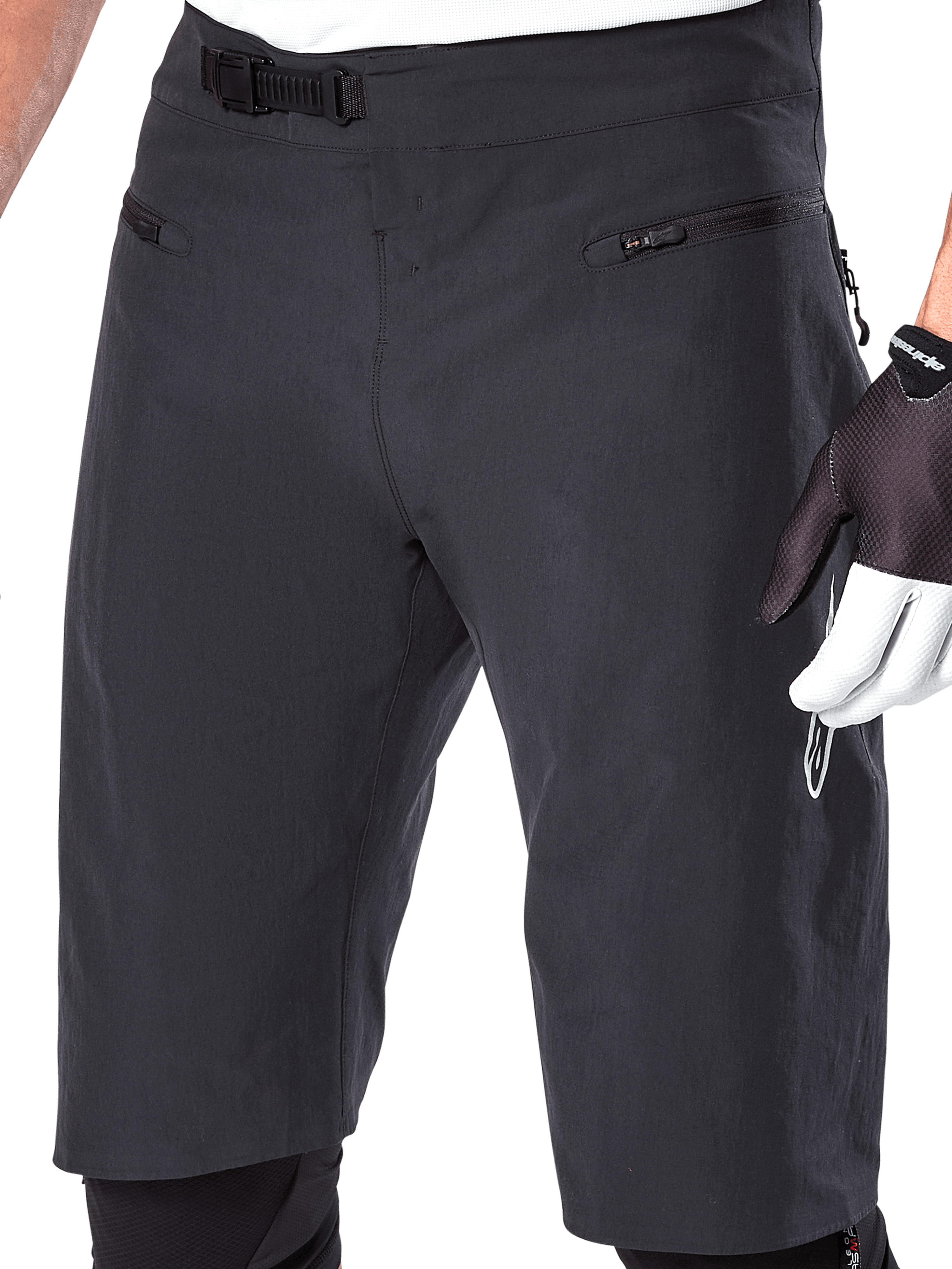 Alpinestars A-Aria Shorts, shorts para ciclismo de montaña, negros, vista de primer plano de la parte frontal de la cintura y los muslos que muestra el sistema de cierre de trinquete y los bolsillos con cremallera, usados con guantes de ciclismo blancos y negros, equipamiento técnico de trail.