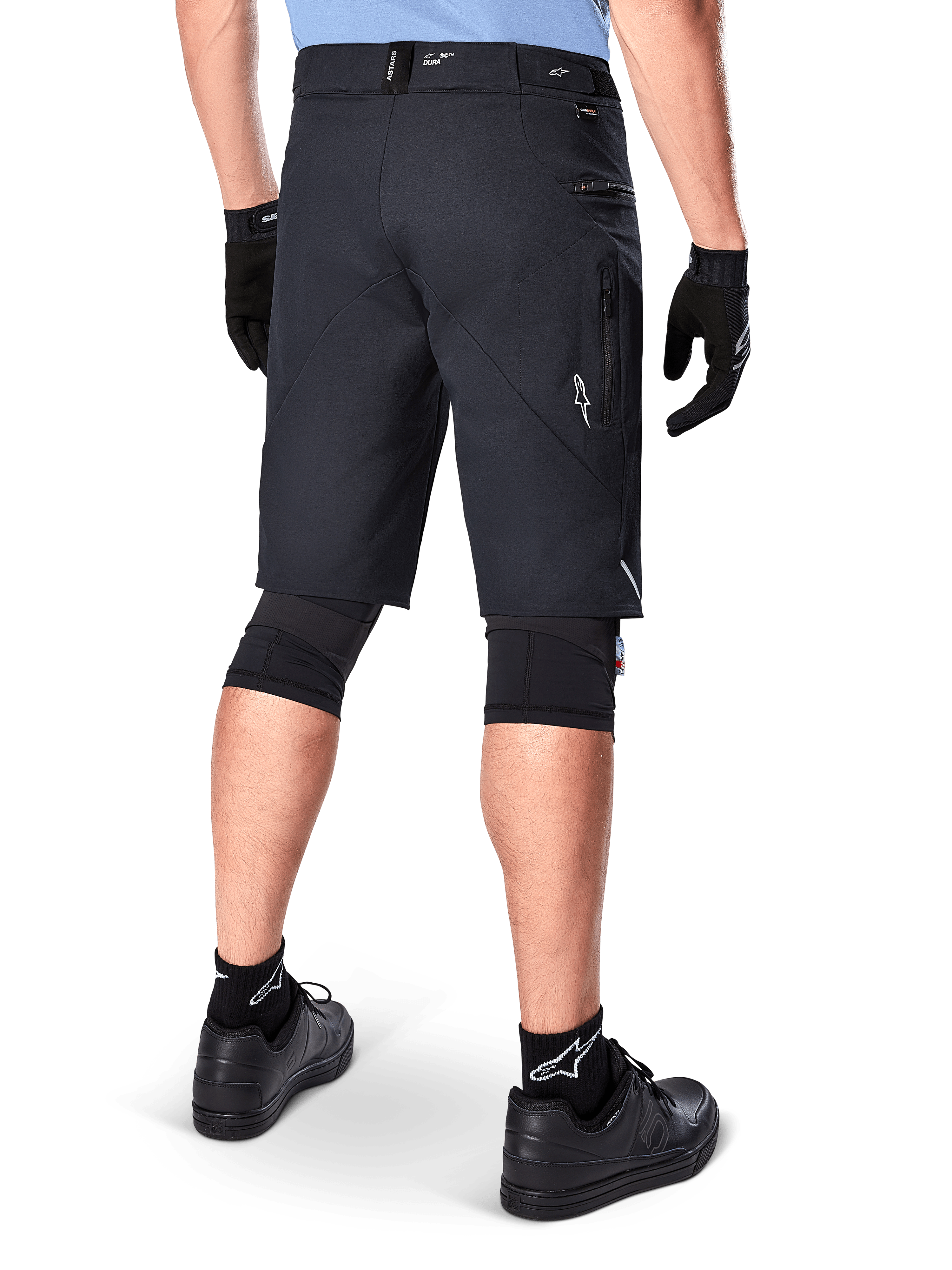 Shorts Alpinestars A-Dura Elite, negros, vista trasera de un ciclista con equipo de bicicleta de montaña que incluye rodilleras negras, guantes y calcetines con logos blancos, con bolsillos laterales con cremallera y detalles reflectantes en un tejido Cordura resistente.