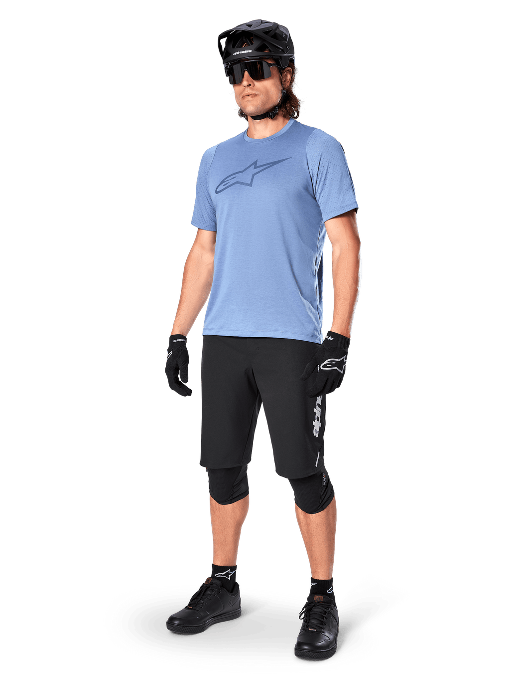 Ciclista con shorts Alpinestars A-Dura Elite en color negro, jersey de manga corta azul claro con logo en el pecho, casco negro, gafas de sol y rodilleras protectoras, equipamiento completo de ciclismo de montaña para rutas todoterreno