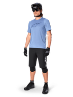 Ciclista con shorts Alpinestars A-Dura Elite en color negro, jersey de manga corta azul claro con logo en el pecho, casco negro, gafas de sol y rodilleras protectoras, equipamiento completo de ciclismo de montaña para rutas todoterreno