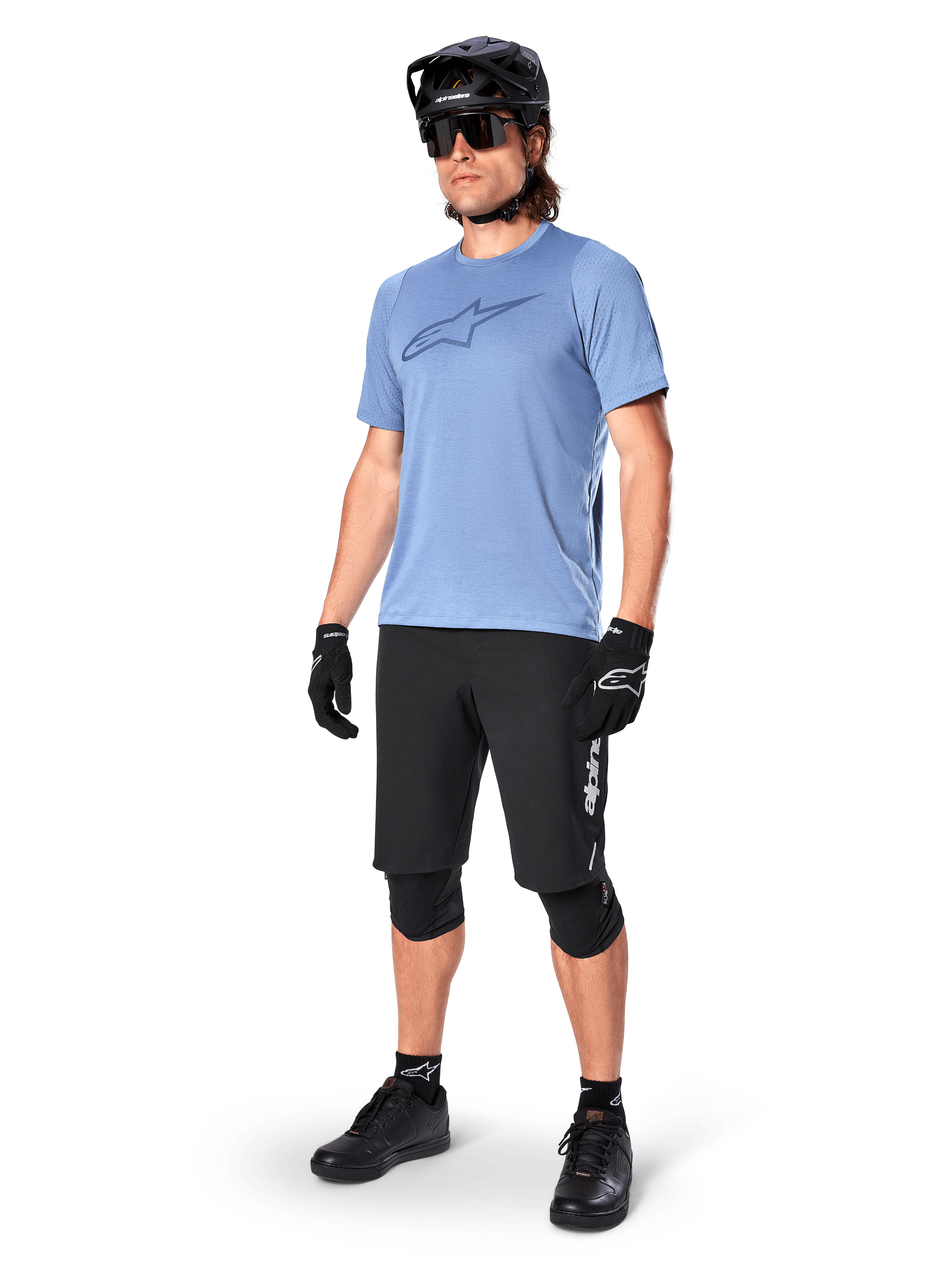 Ciclista con shorts Alpinestars A-Dura Elite en color negro, jersey de manga corta azul claro con logo en el pecho, casco negro, gafas de sol y rodilleras protectoras, equipamiento completo de ciclismo de montaña para rutas todoterreno