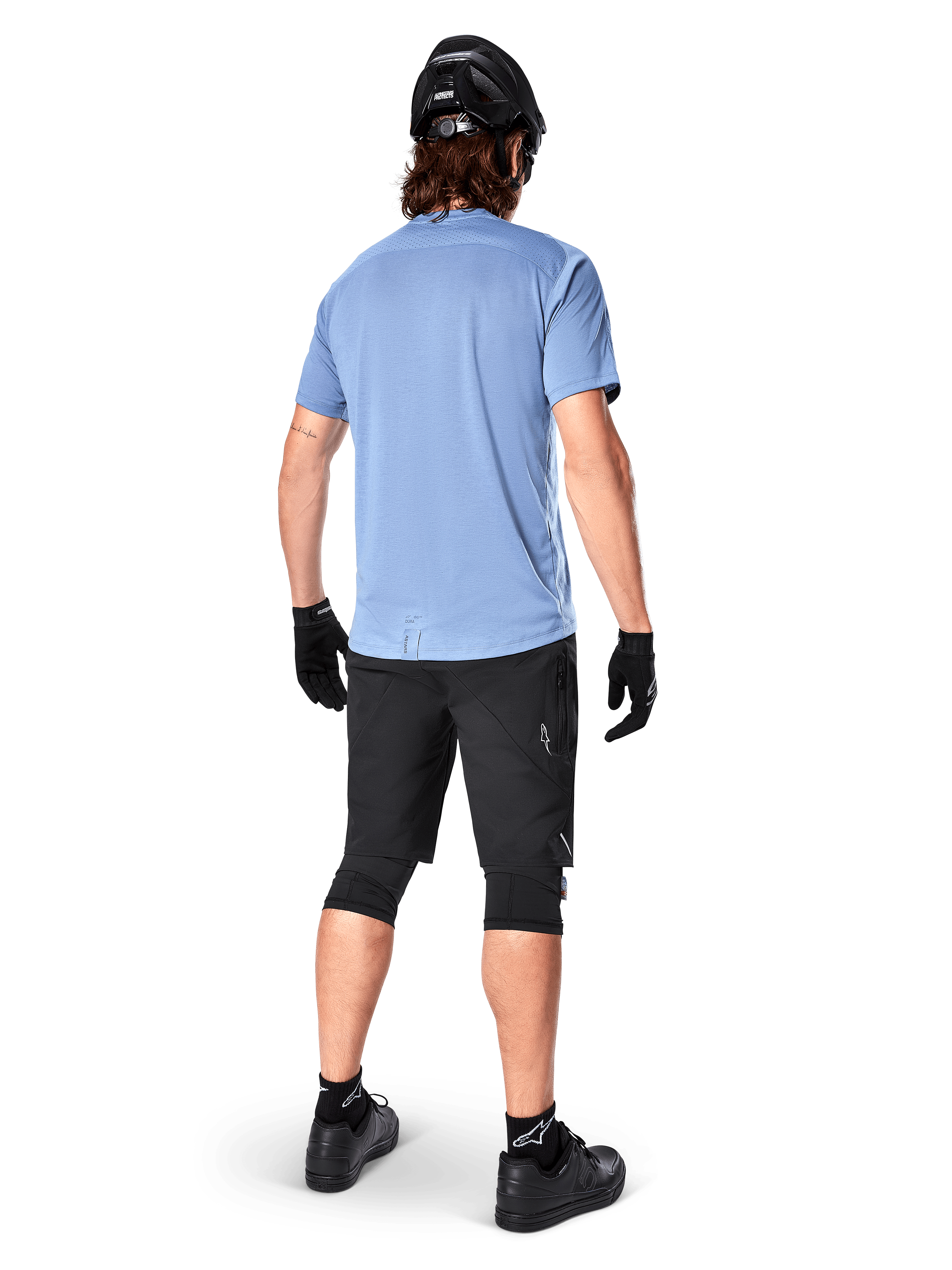 Alpinestars A-Dura Elite Shorts, pantalones cortos de ciclismo de montaña, negros, vista trasera en el modelo, usados con camiseta de manga corta azul claro y equipo de protección negro, diseño técnico con bolsillos con cremallera y detalles reflectantes para ciclismo fuera de carretera