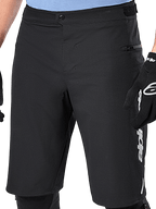 Alpinestars A-Dura Elite Shorts, pantalones cortos de MTB negros, vista de cerca que muestra el cierre frontal de presión y el bolsillo lateral con cremallera, usados por un ciclista con guantes de ciclismo negros y rodilleras, equipamiento de alto rendimiento para ciclismo de montaña