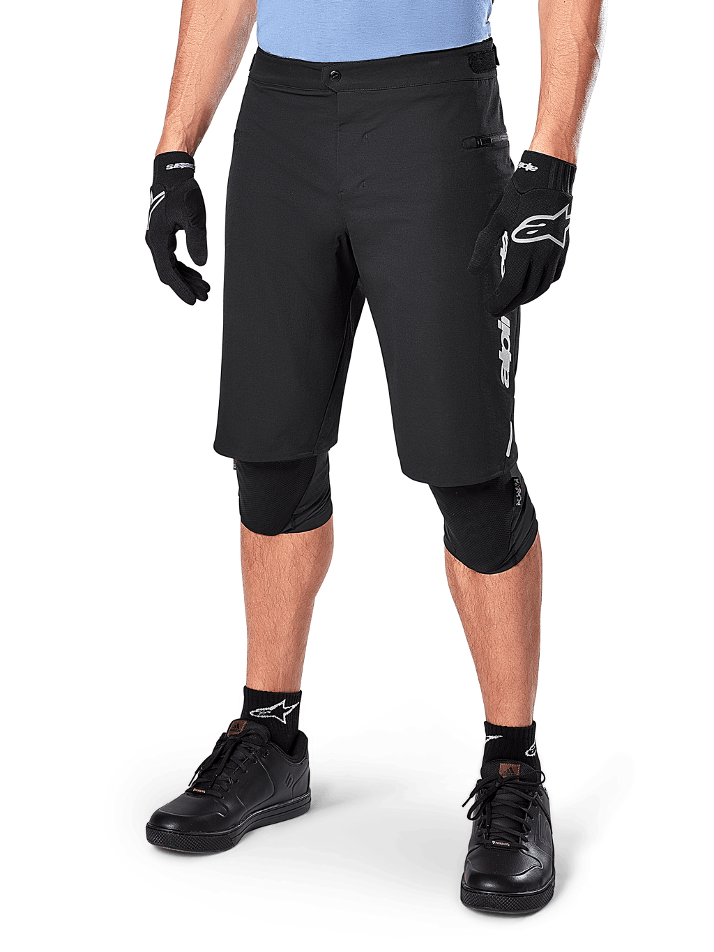 Alpinestars A-Dura Elite Shorts, pantalones cortos negros de ciclismo de montaña (MTB), usados con una camiseta azul claro y protecciones de rodilla negras, con un cierre de presión seguro y bolsillo con cremallera, combinados con zapatillas de ciclismo y guantes negros para un conjunto todoterreno completo.
