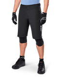 Alpinestars A-Dura Elite Shorts, pantalones cortos negros de ciclismo de montaña (MTB), usados con una camiseta azul claro y protecciones de rodilla negras, con un cierre de presión seguro y bolsillo con cremallera, combinados con zapatillas de ciclismo y guantes negros para un conjunto todoterreno completo.