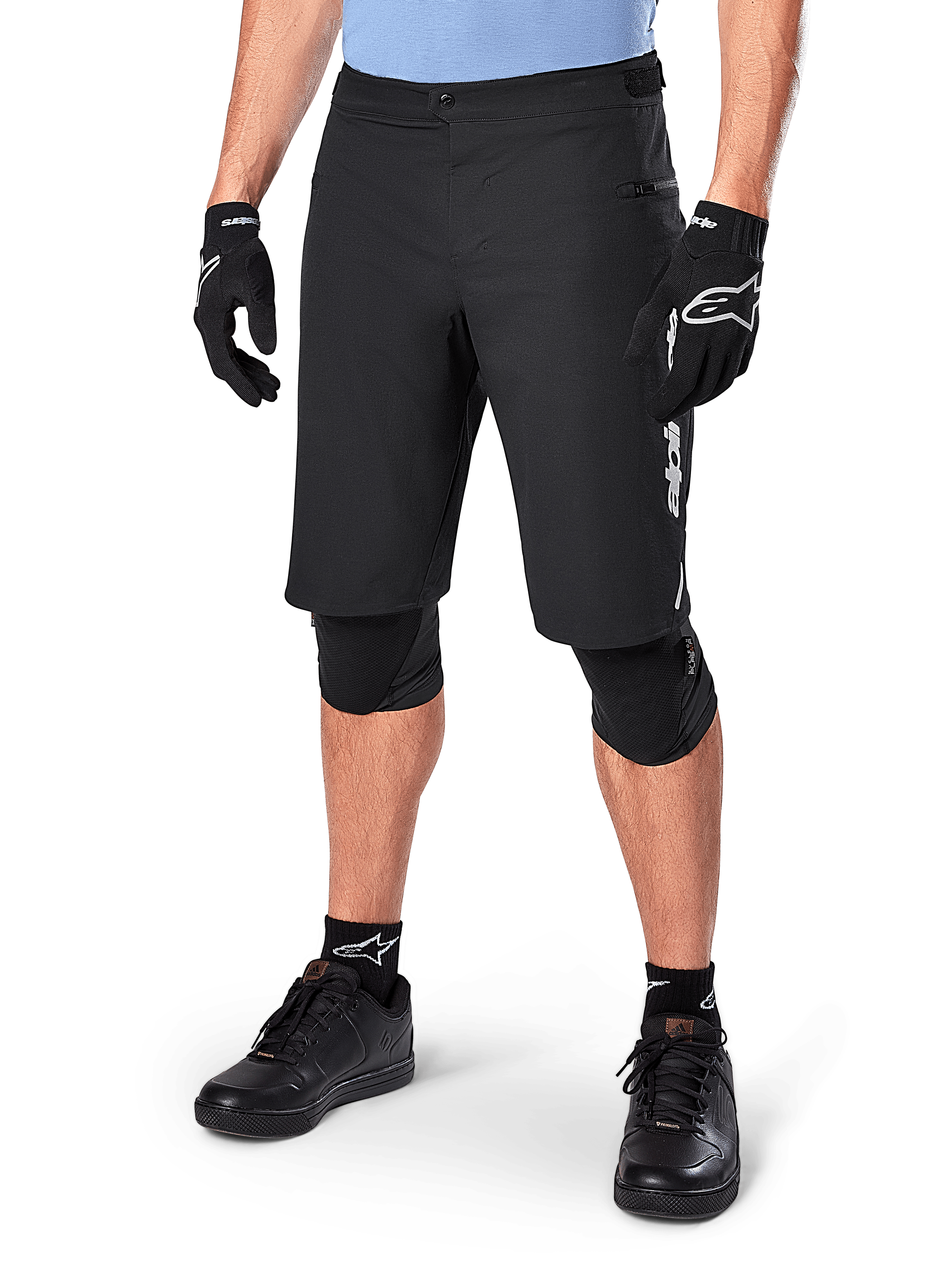 Alpinestars A-Dura Elite Shorts, pantalones cortos negros de ciclismo de montaña (MTB), usados con una camiseta azul claro y protecciones de rodilla negras, con un cierre de presión seguro y bolsillo con cremallera, combinados con zapatillas de ciclismo y guantes negros para un conjunto todoterreno completo.