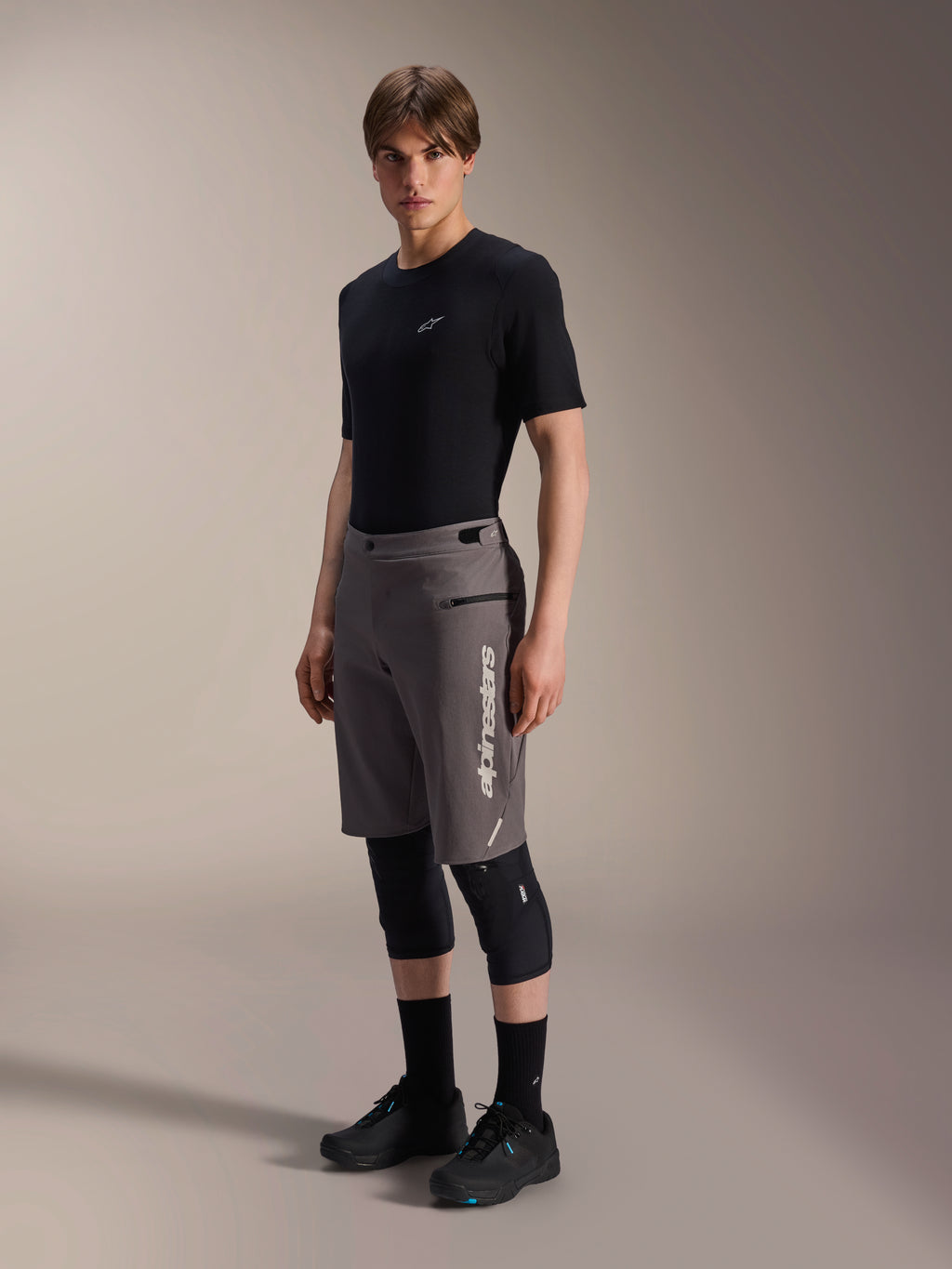 Alpinestars A-Dura Elite Shorts, pantalones cortos de mountain bike gris oscuro, usados por un modelo con camiseta negra y rodilleras, con el logotipo vertical de Alpinestars en blanco en la pierna izquierda, tejido técnico Cordura para ciclismo de montaña.