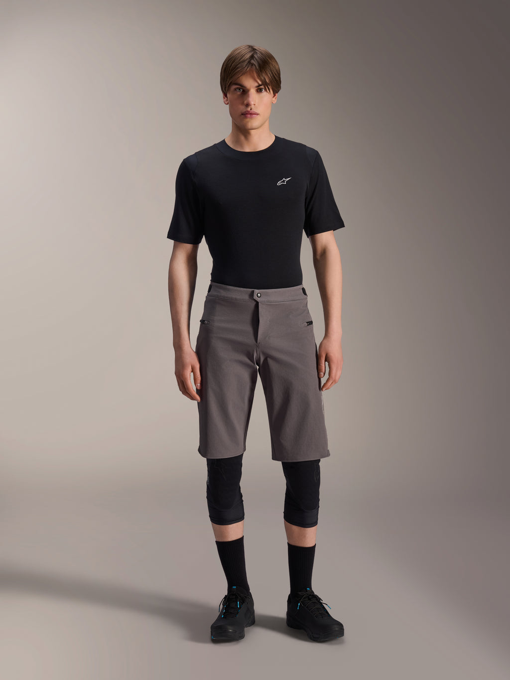 Alpinestars A-Dura Elite Shorts, pantalones cortos de ciclismo de montaña gris oscuro, combinados con una camiseta negra de cuello redondo y rodilleras negras, prenda técnica de longitud hasta la rodilla con cierre frontal a presión y tejido Cordura resistente.