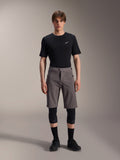 Alpinestars A-Dura Elite Shorts, pantalones cortos de ciclismo de montaña gris oscuro, combinados con una camiseta negra de cuello redondo y rodilleras negras, prenda técnica de longitud hasta la rodilla con cierre frontal a presión y tejido Cordura resistente.