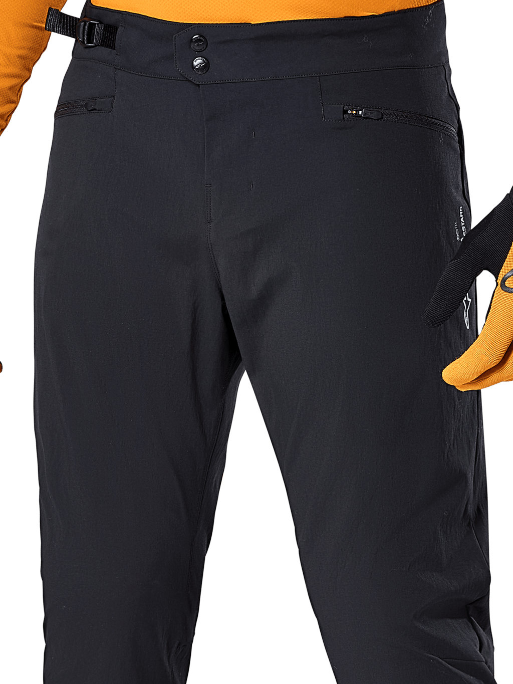 Alpinestars Pantalones A-Dura, pantalones de ciclismo MTB negros, vista de primer plano con cierre de cintura de doble broche, ajustador lateral y bolsillos con cremallera, combinados con un jersey y guantes naranjas