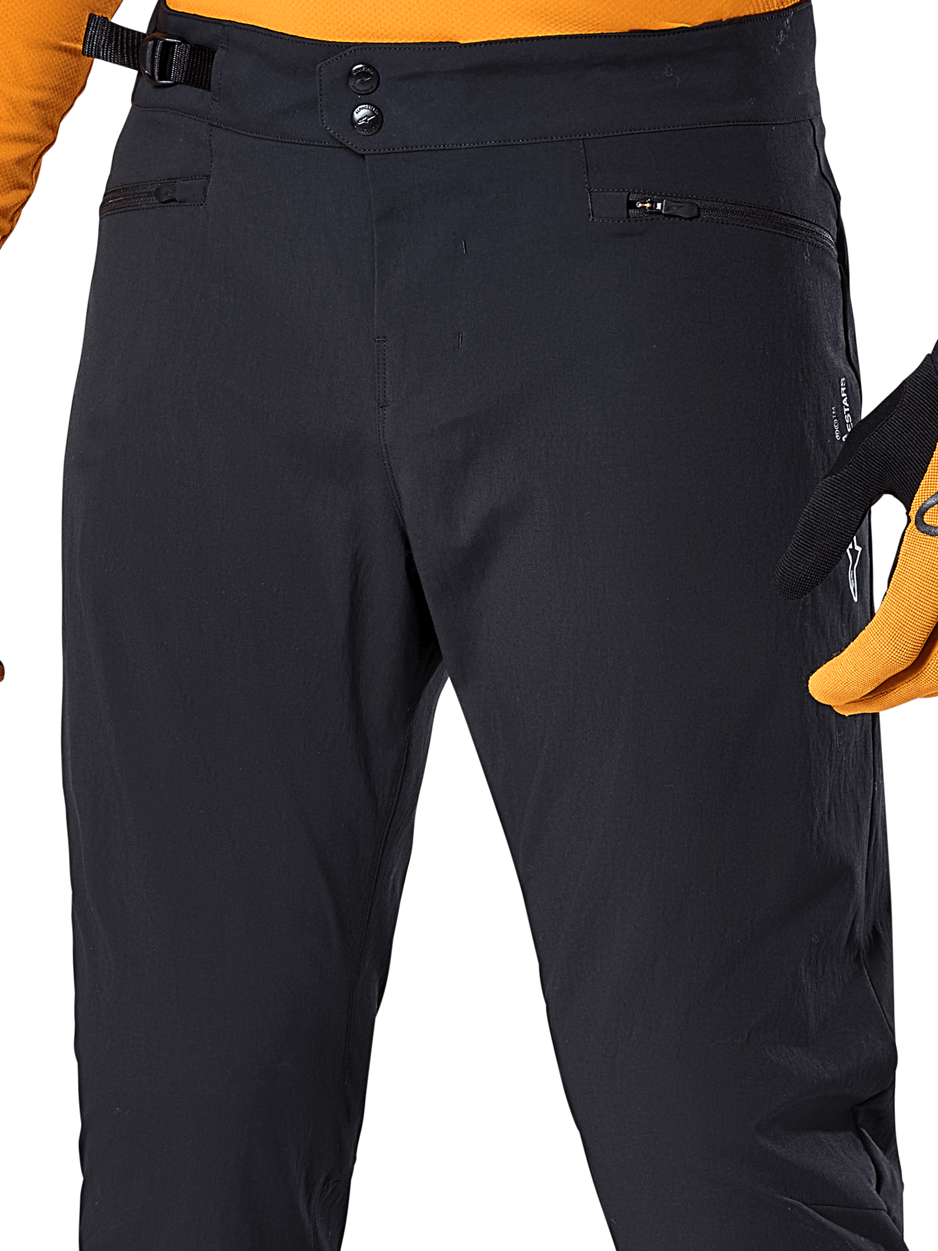 Alpinestars Pantalones A-Dura, pantalones de ciclismo MTB negros, vista de primer plano con cierre de cintura de doble broche, ajustador lateral y bolsillos con cremallera, combinados con un jersey y guantes naranjas