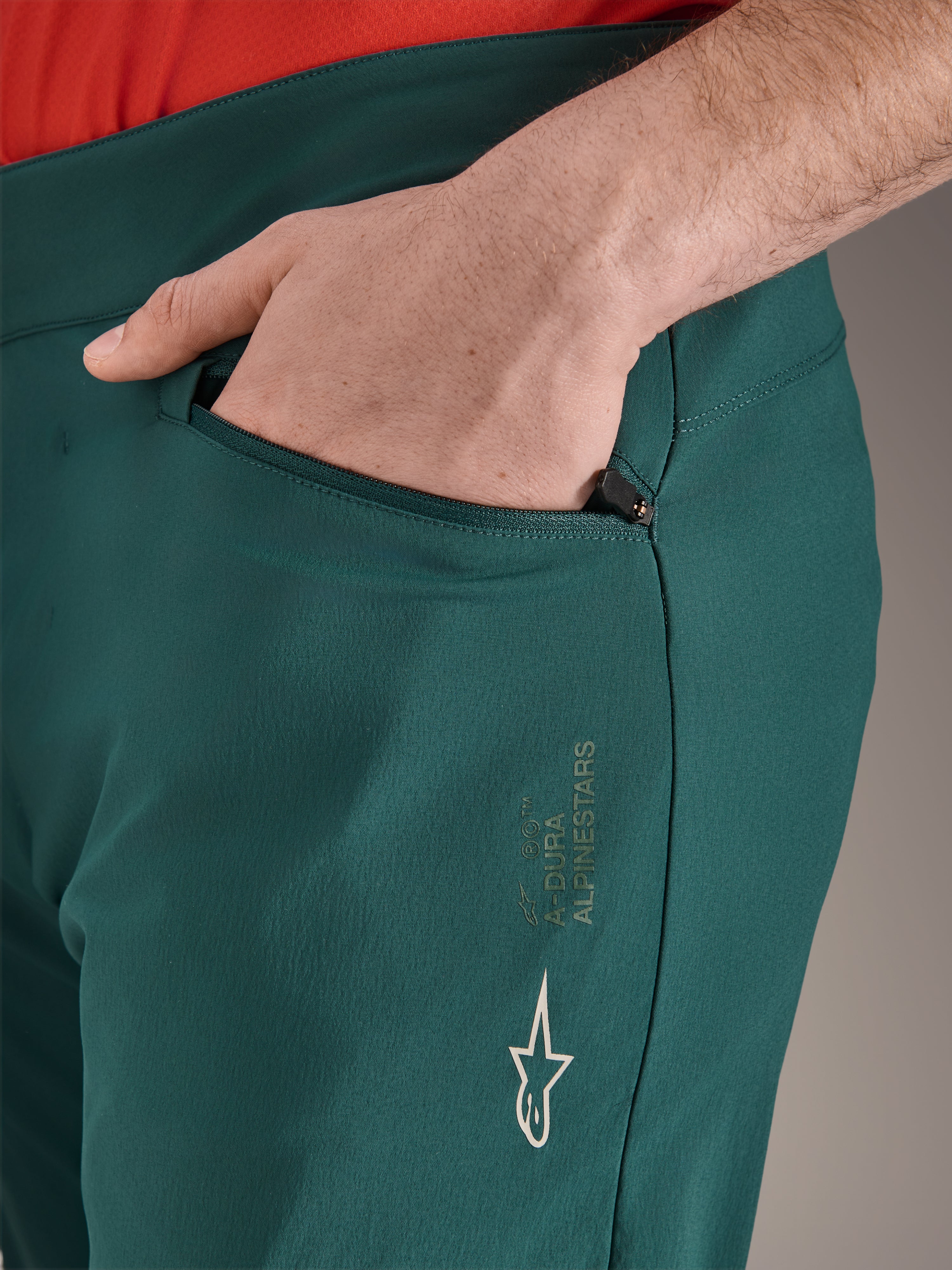 Alpinestars A-Dura Pants in Dark Pine green, close-up of zippered pocket with hand, featuring white Alpinestars logo and text branding on the thigh, durable MTB cycling trousers