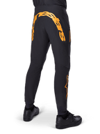 Alpinestars A-Supra Race Pants, pantalones de MTB negros, vista trasera con una gran marca denominativa Alpinestars en naranja en el canesú trasero, logotipo naranja en la parte inferior de la pierna, ajuste de alto rendimiento con bolsillo con cremallera, usados con zapatillas negras