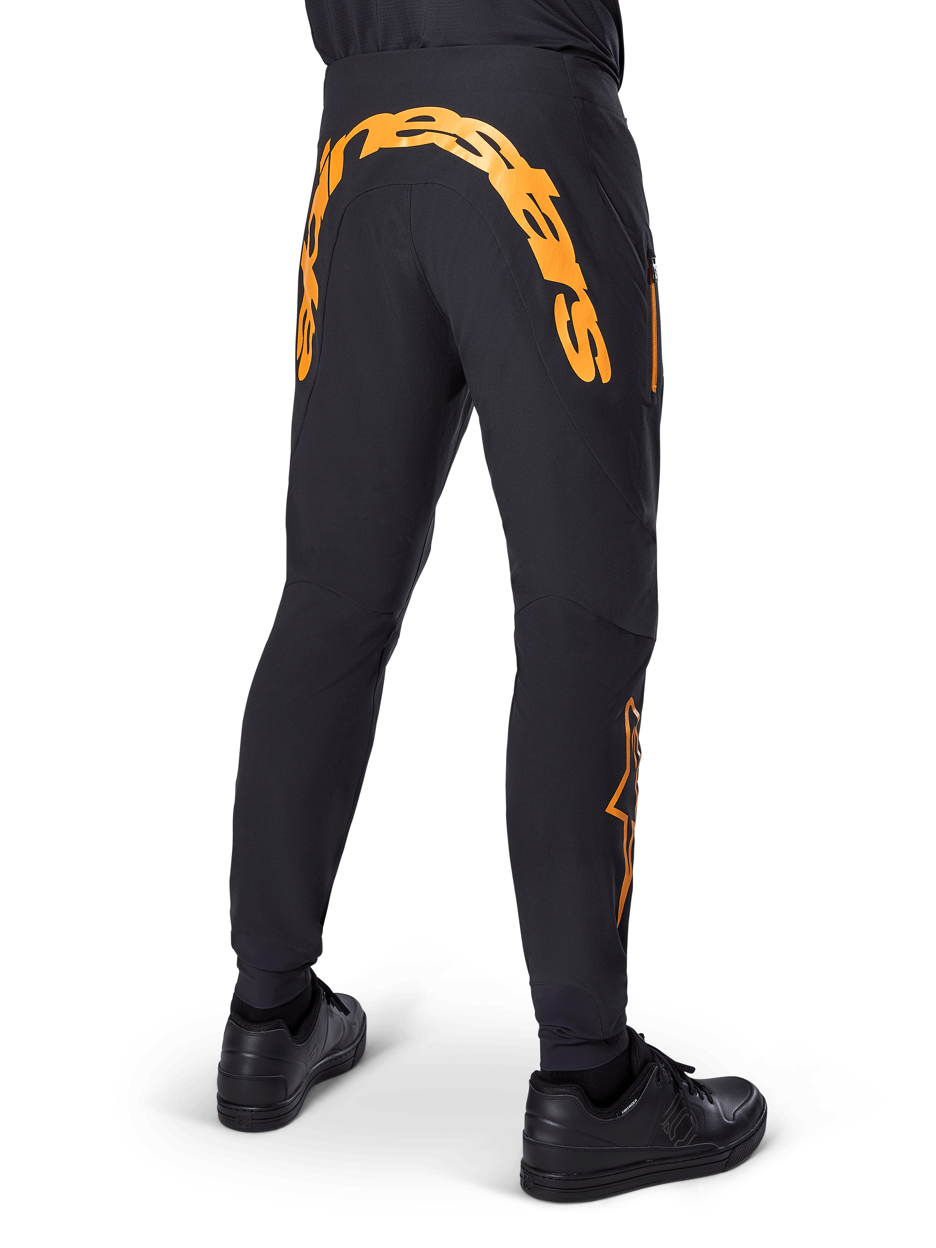 Alpinestars A-Supra Race Pants, pantalones de MTB negros, vista trasera con una gran marca denominativa Alpinestars en naranja en el canesú trasero, logotipo naranja en la parte inferior de la pierna, ajuste de alto rendimiento con bolsillo con cremallera, usados con zapatillas negras
