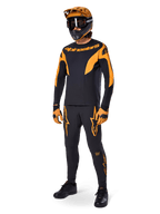 Ciclista de montaña con equipamiento completo de competición, pantalones Alpinestars A-Supra Race en negro con detalles naranjas, jersey de manga larga negro y naranja a juego, casco naranja con gafas negras, guantes naranjas y negros y calzado negro, diseño de kit de competición profesional de MTB
