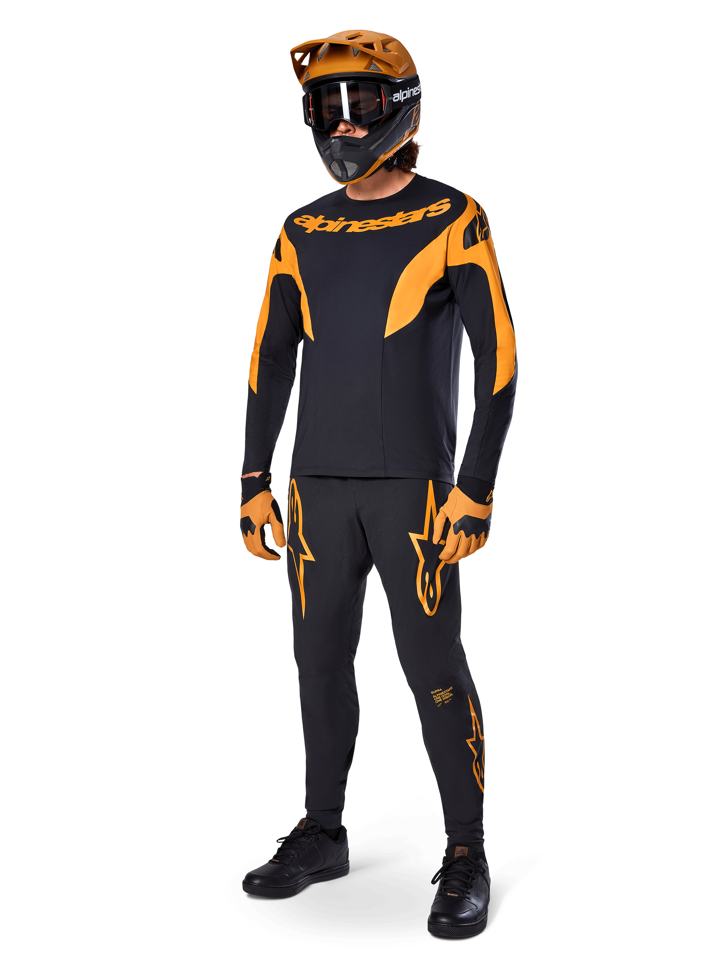 Ciclista de montaña con equipamiento completo de competición, pantalones Alpinestars A-Supra Race en negro con detalles naranjas, jersey de manga larga negro y naranja a juego, casco naranja con gafas negras, guantes naranjas y negros y calzado negro, diseño de kit de competición profesional de MTB