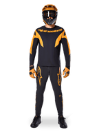 Pantalones Alpinestars A-Supra Race, pantalones de ciclismo de montaña, negros y naranjas, usados por un piloto con un equipamiento de carreras completo que incluye jersey negro y naranja, casco naranja y guantes a juego, con llamativos logotipos de estrella naranjas en las piernas